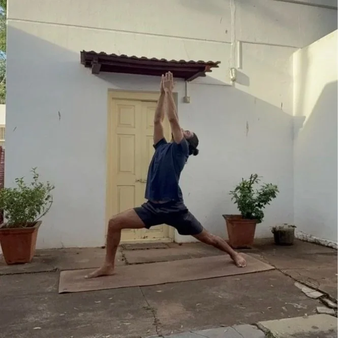Person practicing yoga outdoors on a mat in front of a white wall with a door, plants in pots, performing a warrior pose with hands raised overhead.