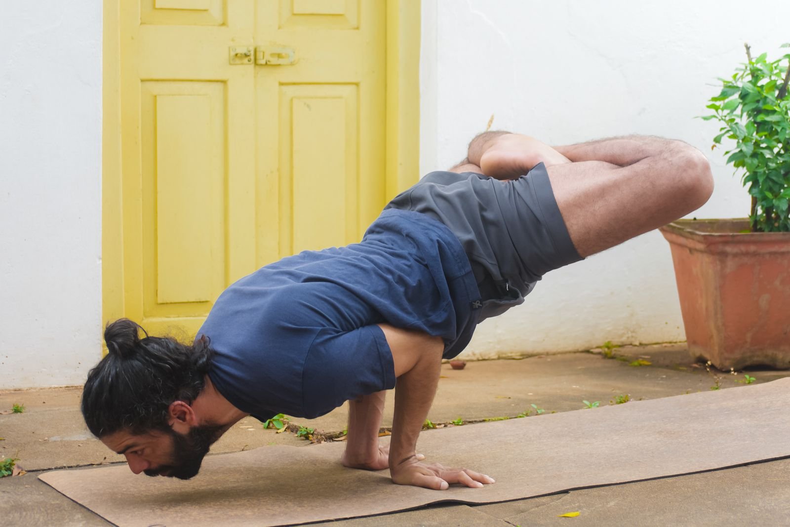 A man with long hair and a beard practicing yoga on a mat outdoors, balancing in a forearm stand position with legs extended back, near a yellow door and potted plant.