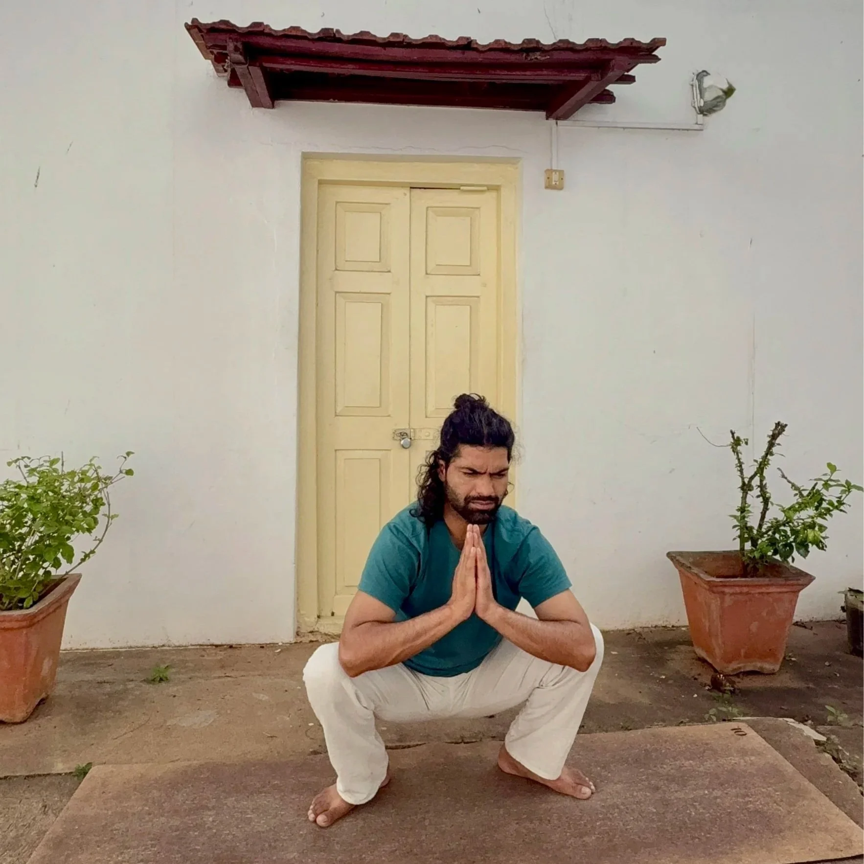 A man practicing yoga outdoors, sitting in a squat with hands pressed together in a prayer position, surrounded by potted plants, with a closed yellow door and a small roof above.