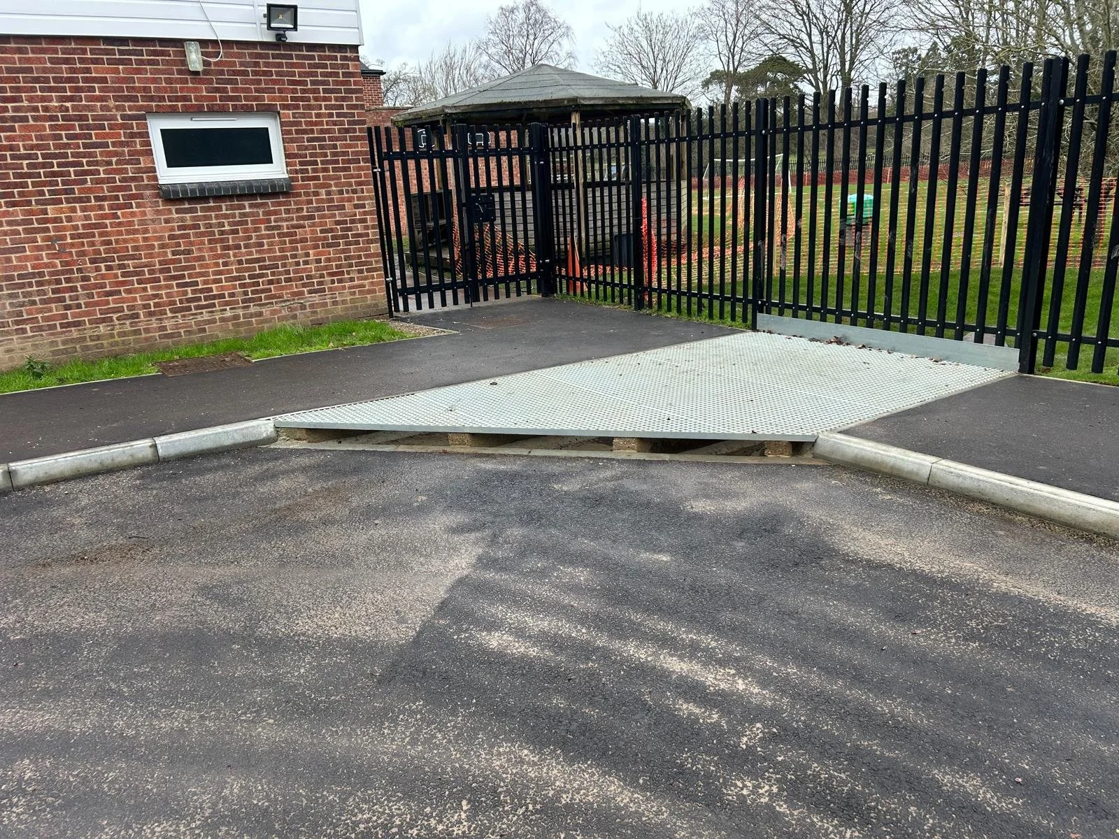 A small ramp for wheelchair access in a parking lot next to a brick building with a white-framed window and a black metal fence surrounding a grassy area with trees.