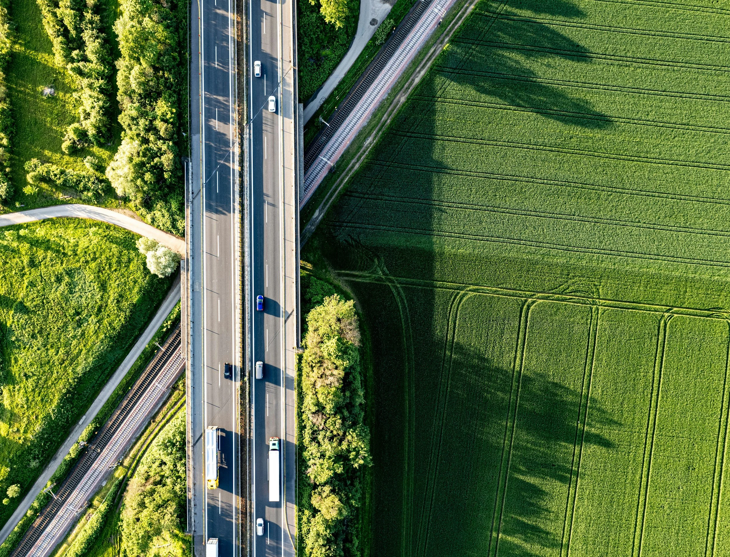 An aerial view of a highway with cars and trucks, surrounded by green fields and trees, crossing over a railway track.