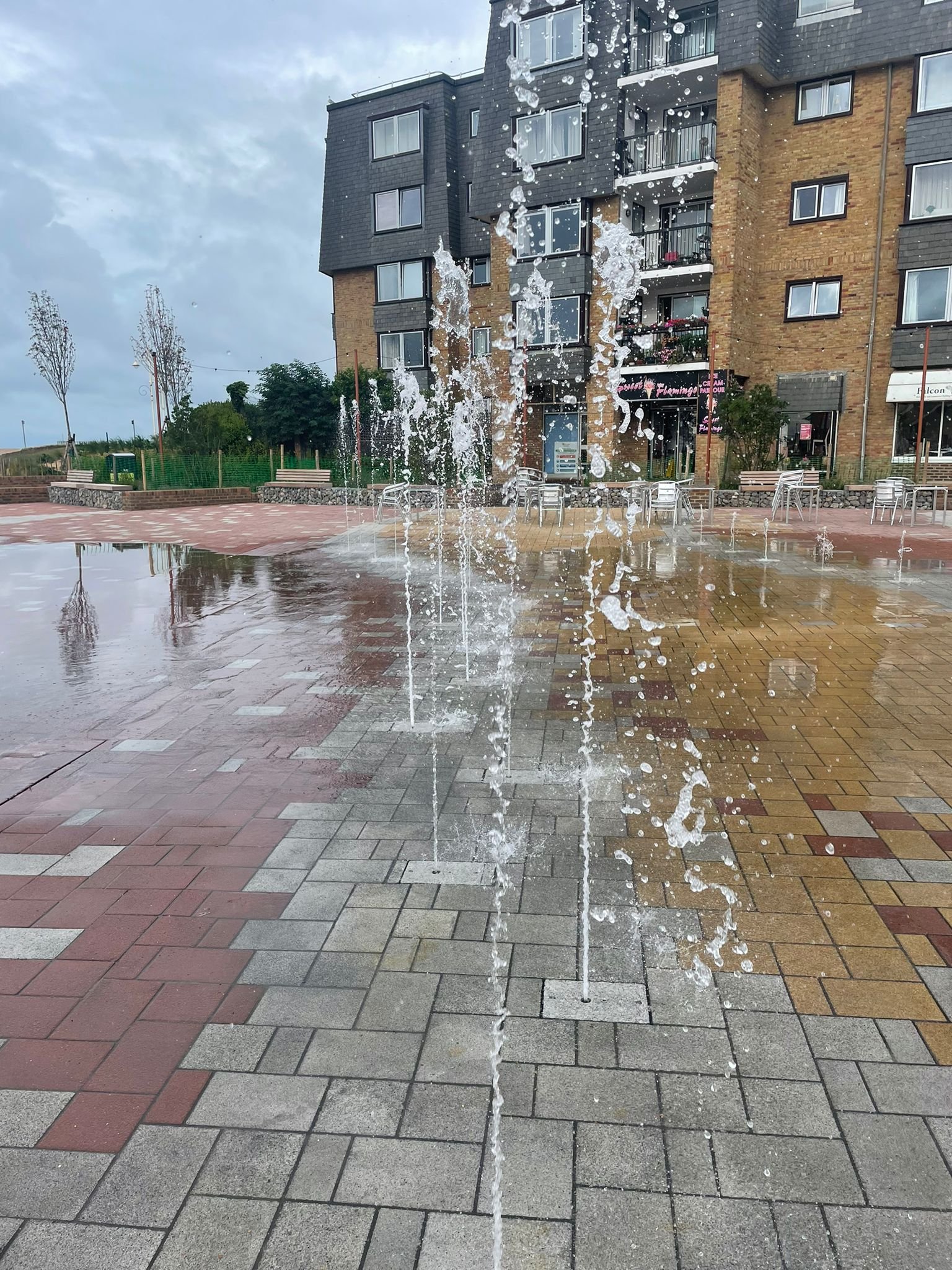 Water fountain with multiple streams of water in an outdoor plaza area with brick and tile paving. In the background, there is a modern multi-story apartment building with balconies and large windows. Overcast sky with some clouds.