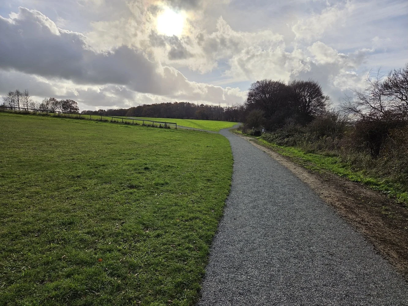A winding gravel path through a grassy field with trees on the right and a cloudy sky with the sun shining.
