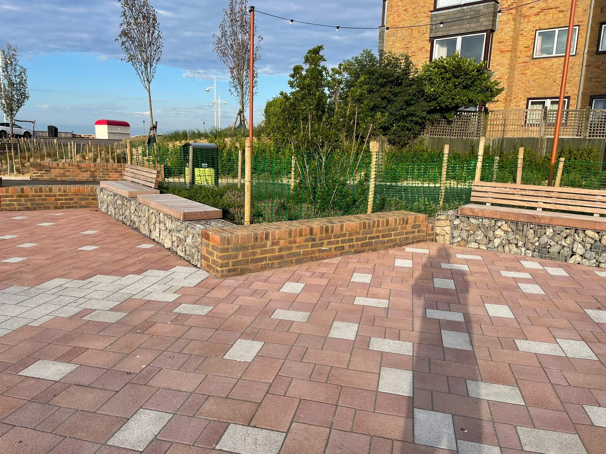 A brick-paved outdoor seating area near a building with trees, benches, a trash can, and string lights.