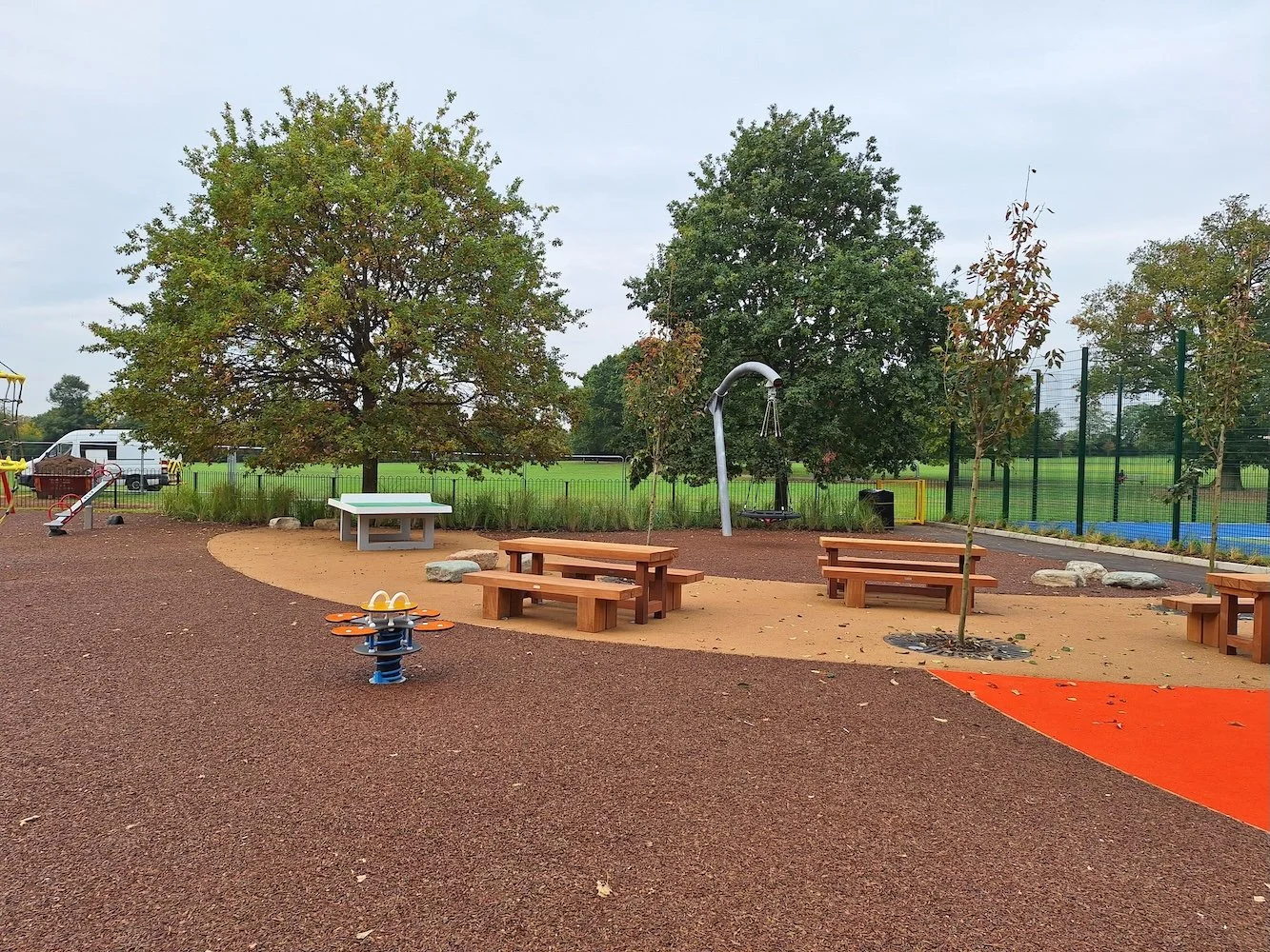 A spacious playground with picnic tables, a swing, a spring rider, and children's play equipment, surrounded by trees and a fenced sports area.