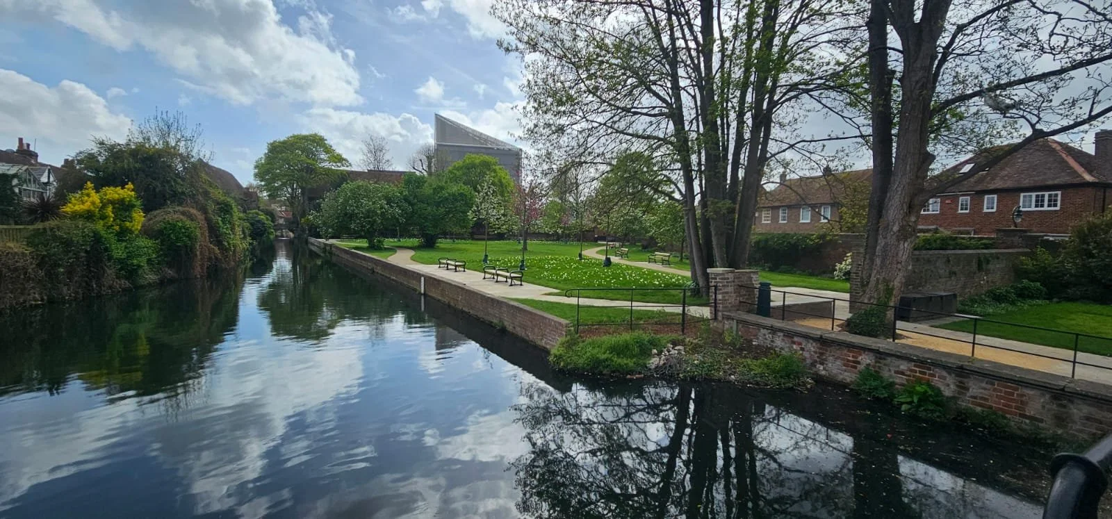 A peaceful park with a river running alongside a walking path. There are benches, trees, and well-maintained grass and shrubs, with buildings visible in the background and a partly cloudy sky.