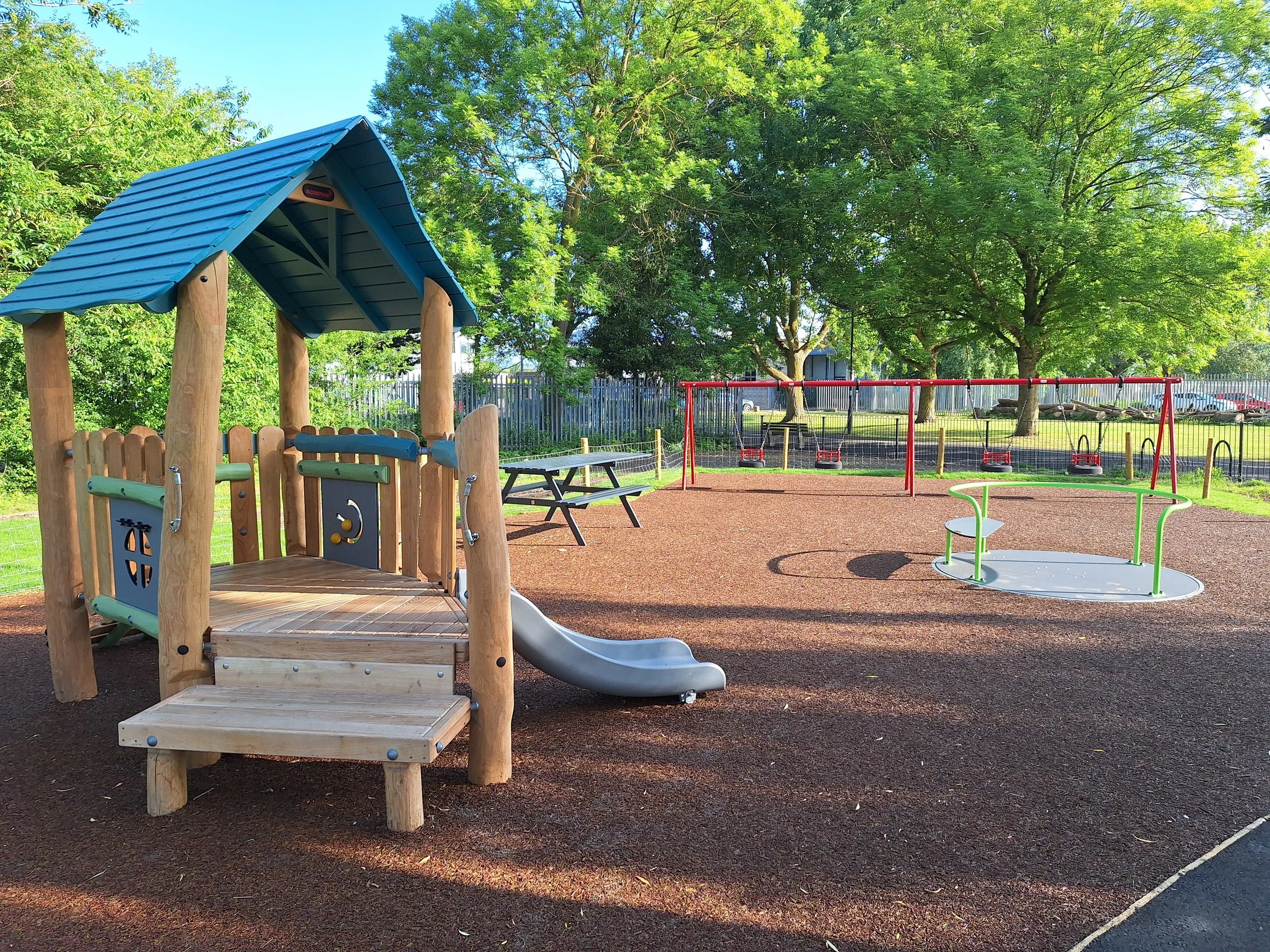 A playground with a shaded wooden play structure with a slide, surrounded by trees, a picnic table, and swings, on a sunny day.