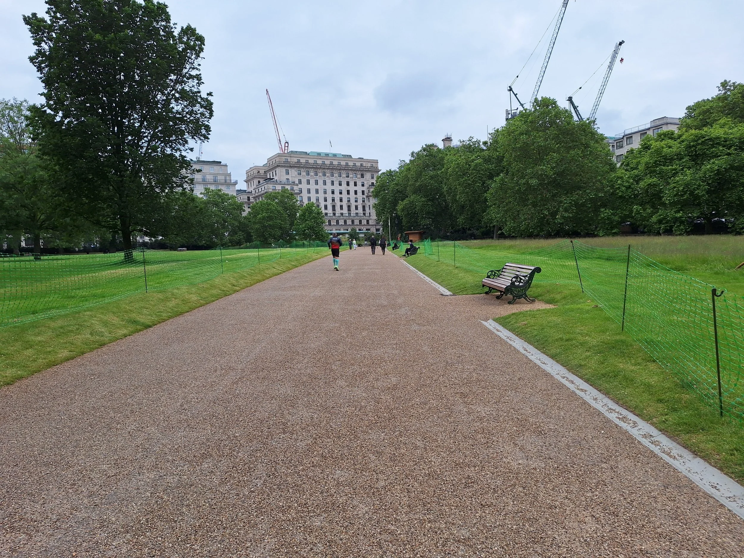 A park pathway with benches and green fencing on both sides, tall trees, and buildings with construction cranes in the background, under a cloudy sky.