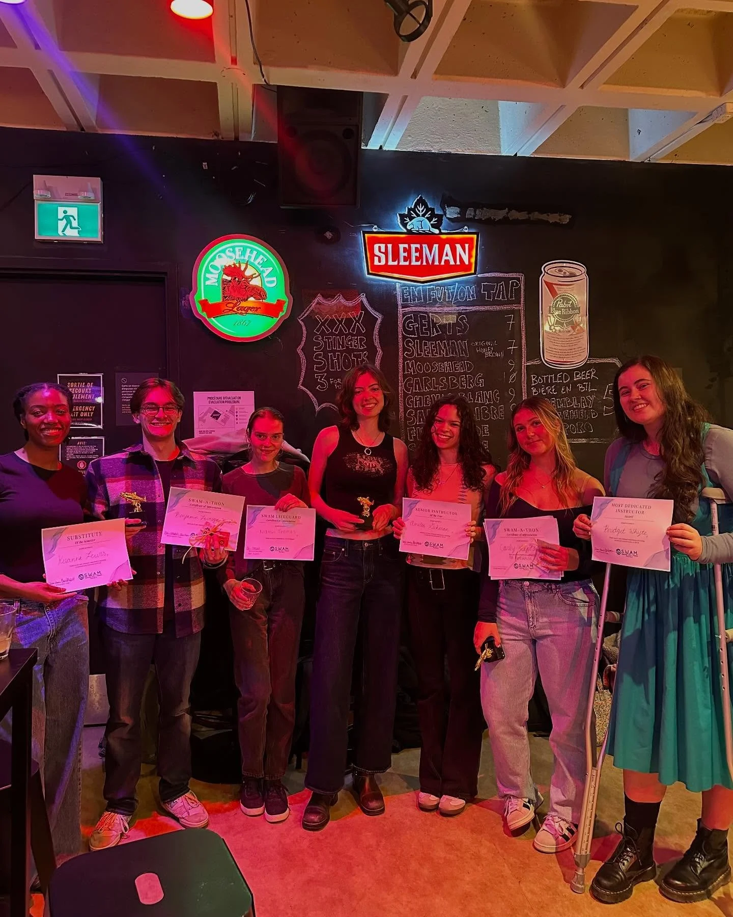 volunteers standing inside a bar, holding certificates, with neon signs and a chalkboard menu behind them