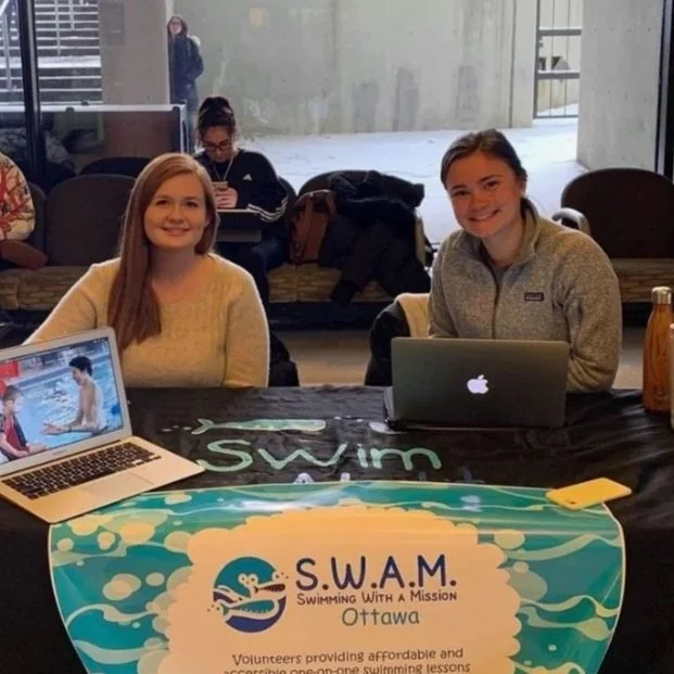 two volunteers sitting at a table with laptops, smiling at the camera, promoting Swimming With A Mission Ottawa.