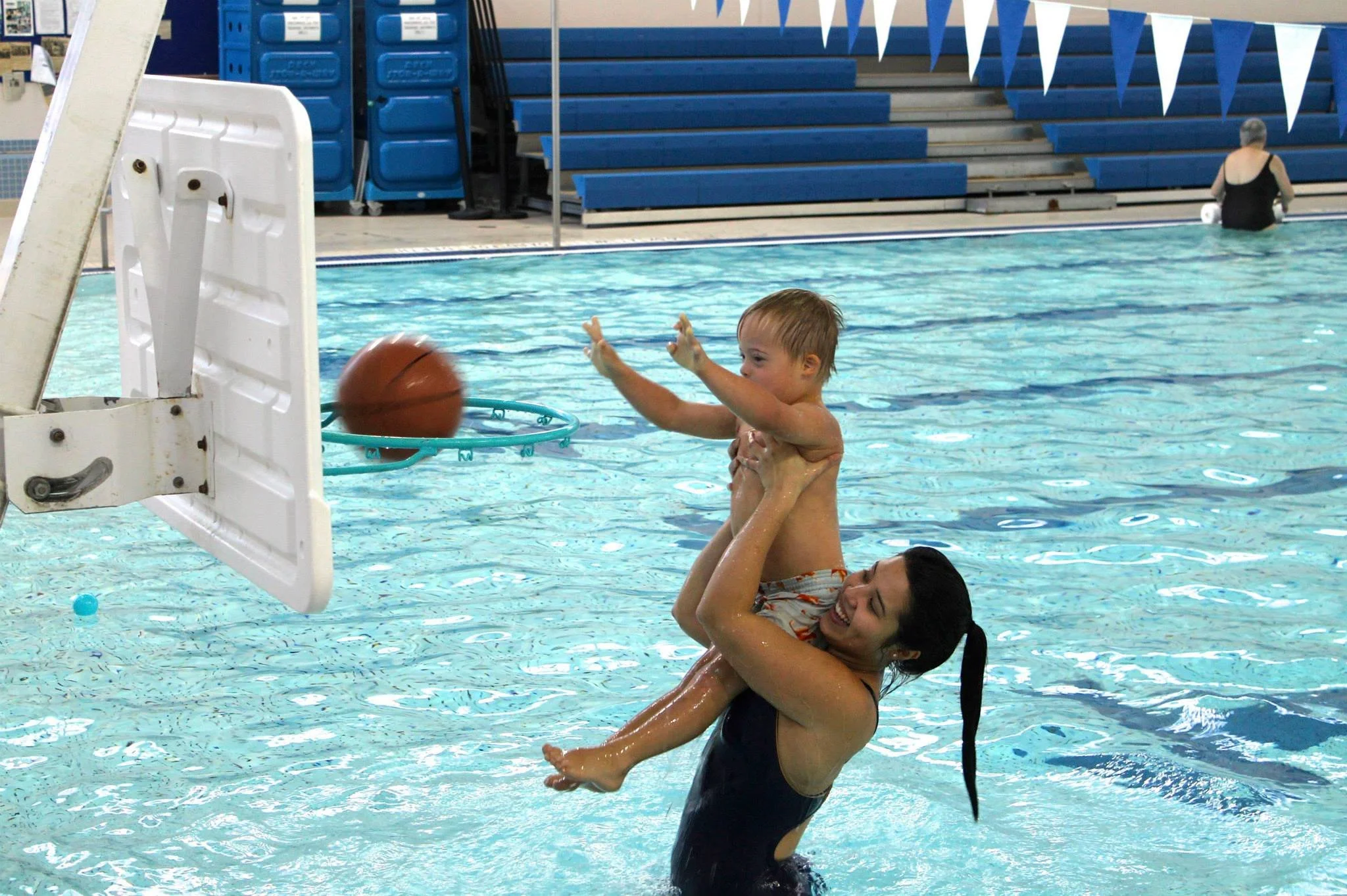 SWAM instructor helping a swimmer shoot a basketball in the hoop, from inside the water