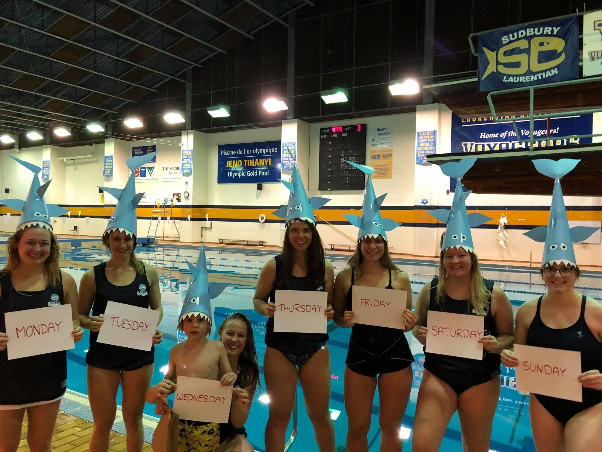 a group of people wearing shark hats and swimming attire, standing by a pool, holding signs with the days of the week written on them