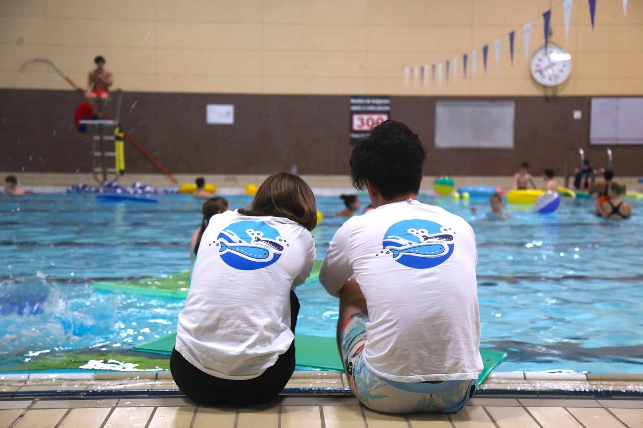 two SWAM instructors sitting by the edge of the pool watching lessons, with SWAM logos on the back of their shirts