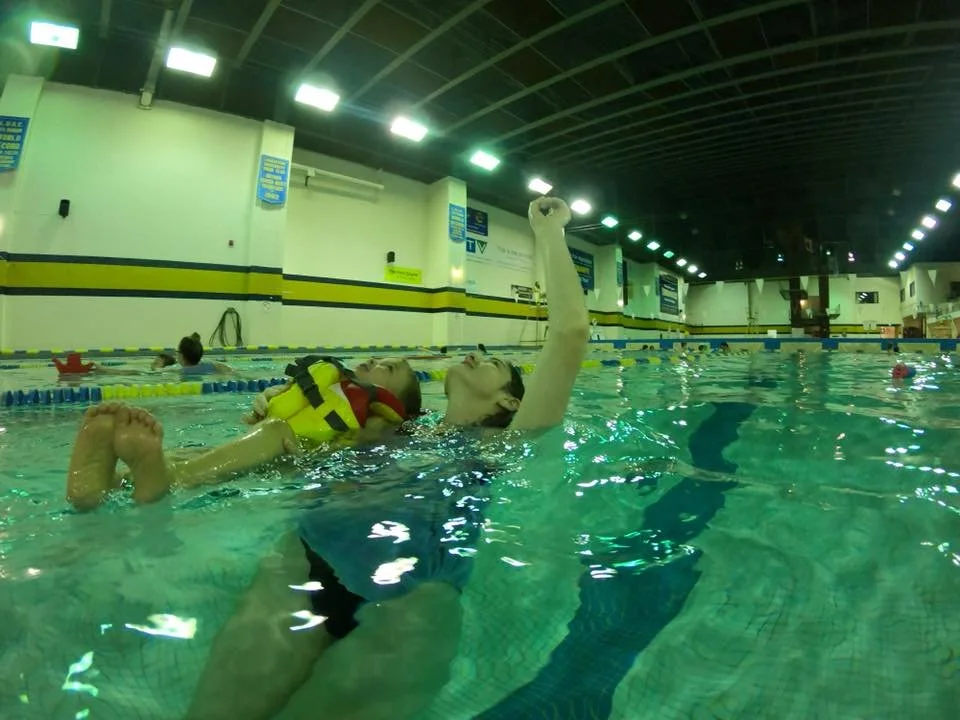 swimmer, wearing lifejacket, and instructor float on their backs and look up at the sky