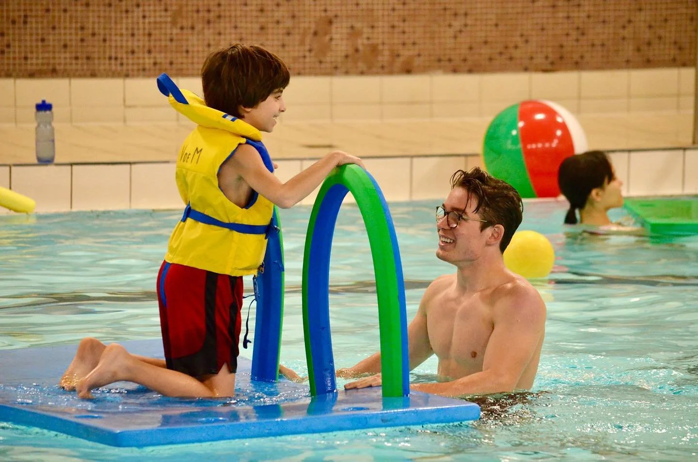 swimmer on floating mat with two noodles smiling at instructor who is in the water