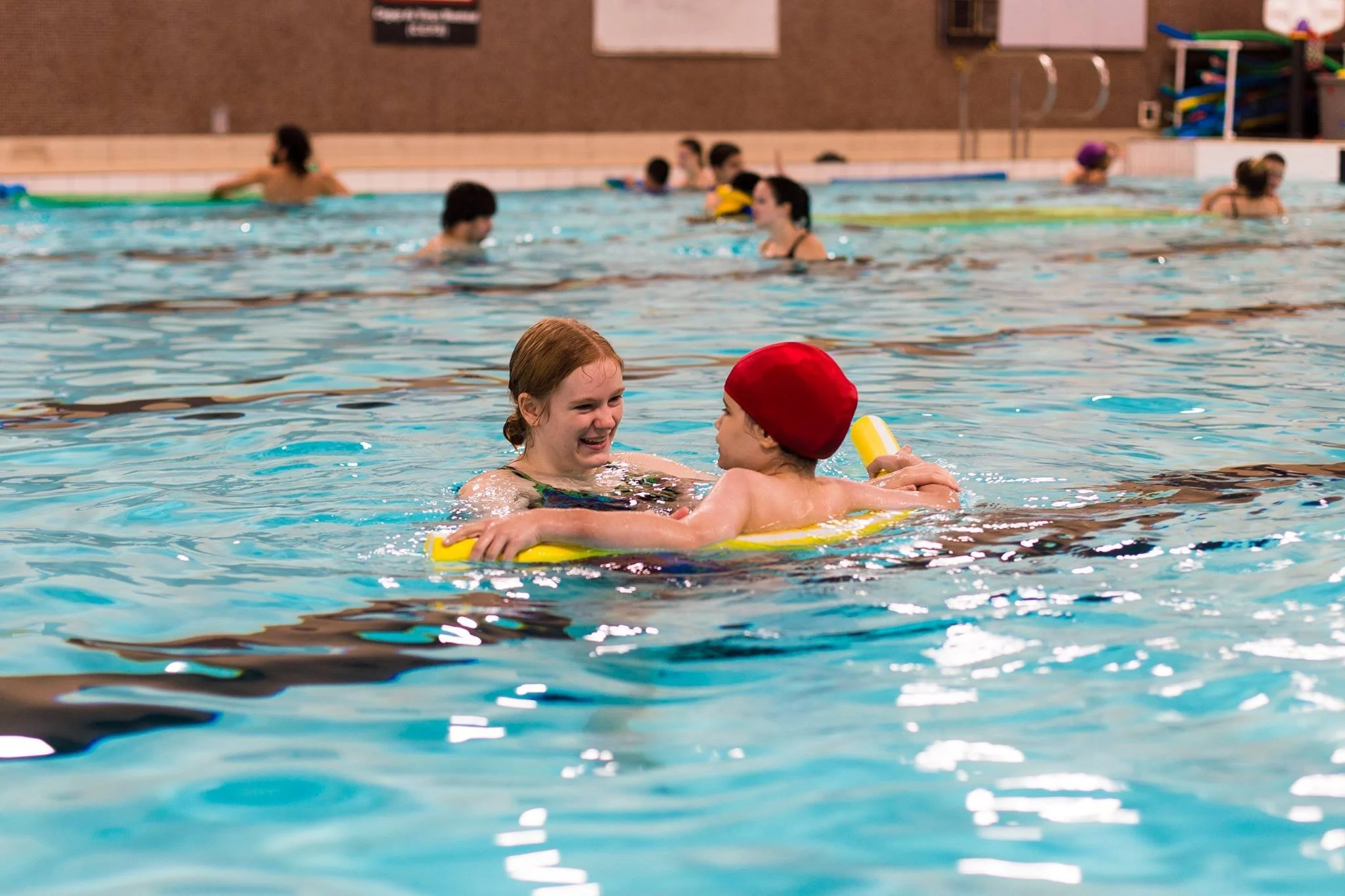 swimmer with red swim cap using noodle, looking at smiling instructor