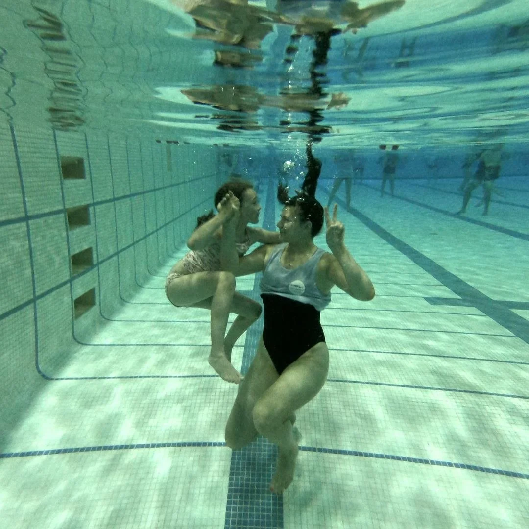 swimmer-instructor pair doing peace signs underwater, looking at each other