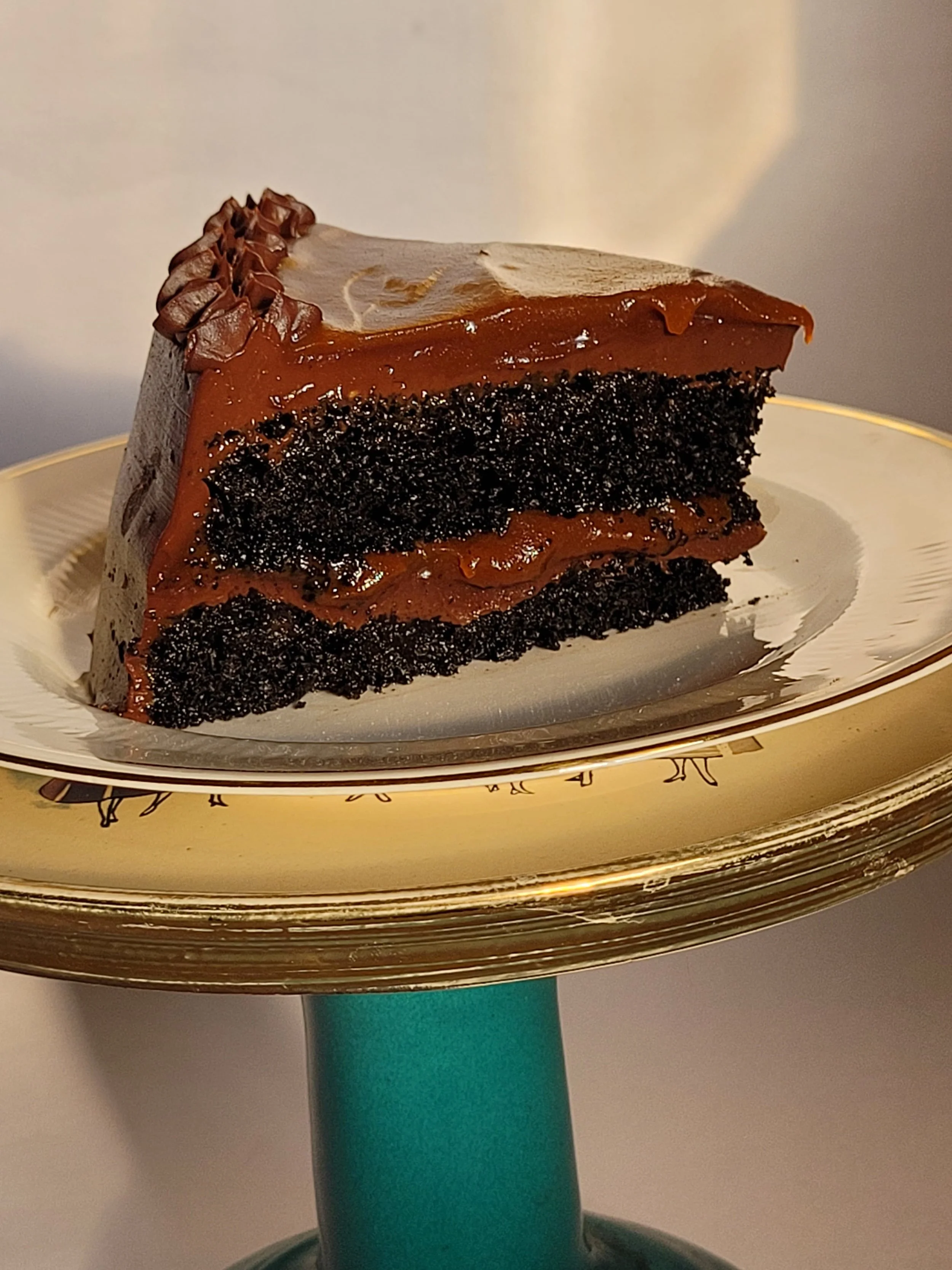 A slice of chocolate cake with dark chocolate layers, caramel frosting, and chocolate sprinkles on top, served on a decorative plate.