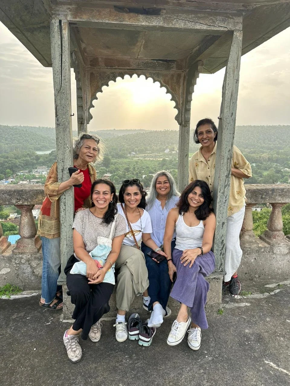 Six women smiling on a viewpoint with a stone structure and scenic landscape in the background, during sunset.