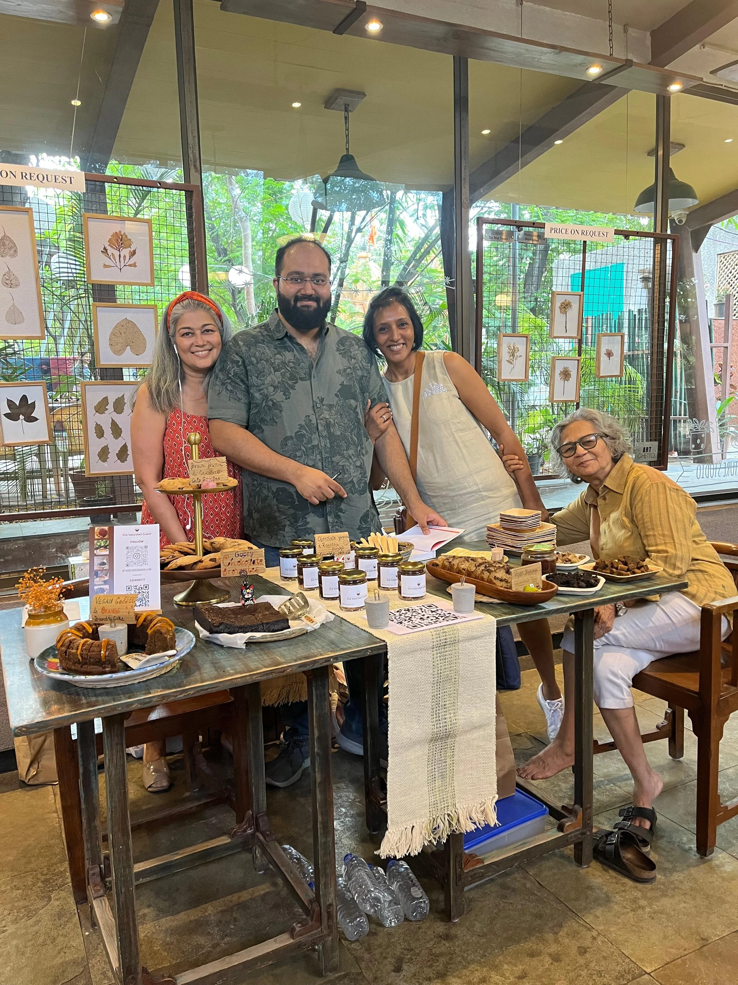 Group of four women and one man smiling behind a table of baked goods, jars, and plates in a bakery or cafe setting with artwork of leaves on the window behind them.