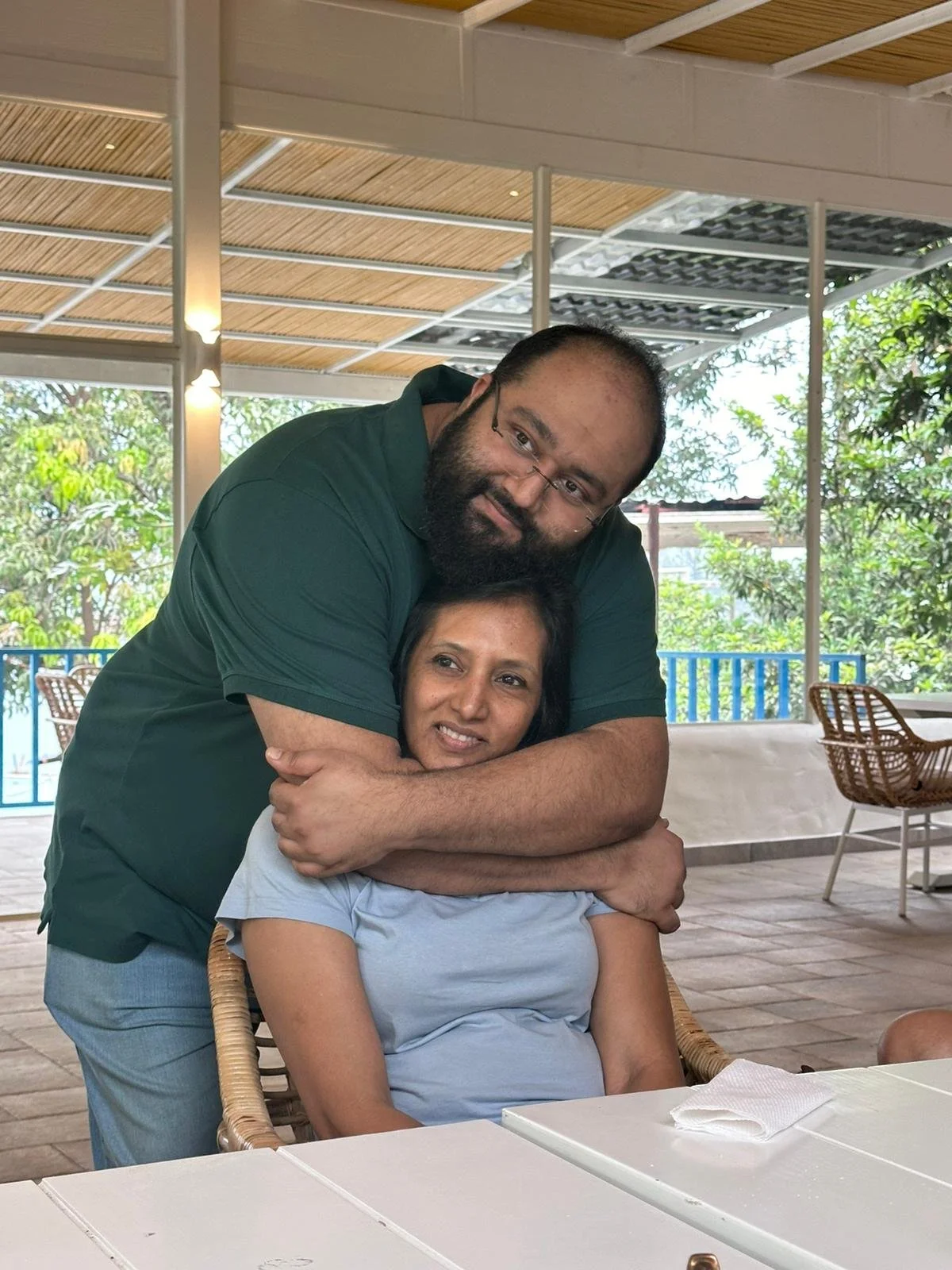 A man with glasses and a beard is hugging a woman in a light blue shirt from behind at a restaurant with a tropical background.