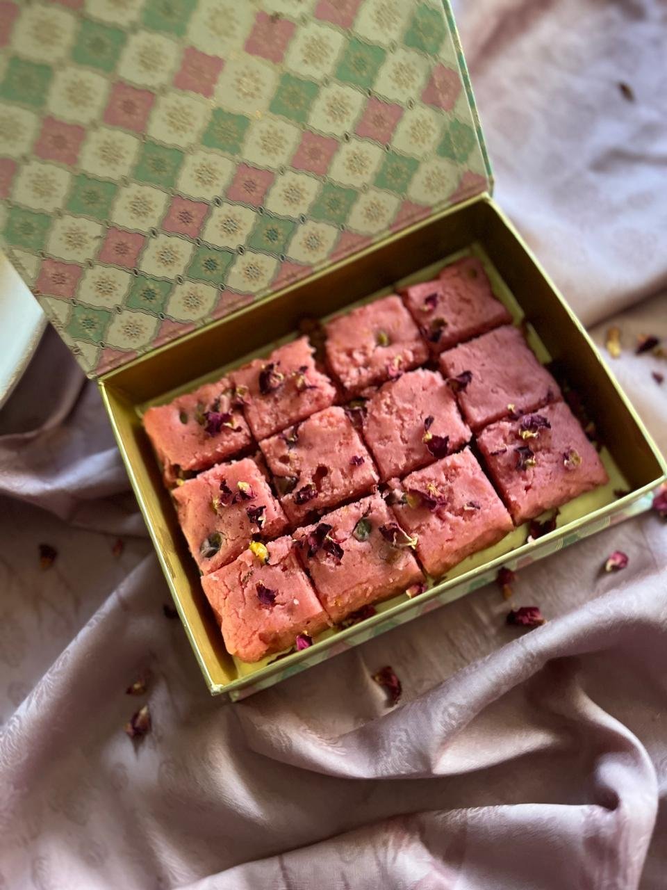 A decorative box containing rose brownie squares topped with dried rose petals, placed on a satin fabric surface.