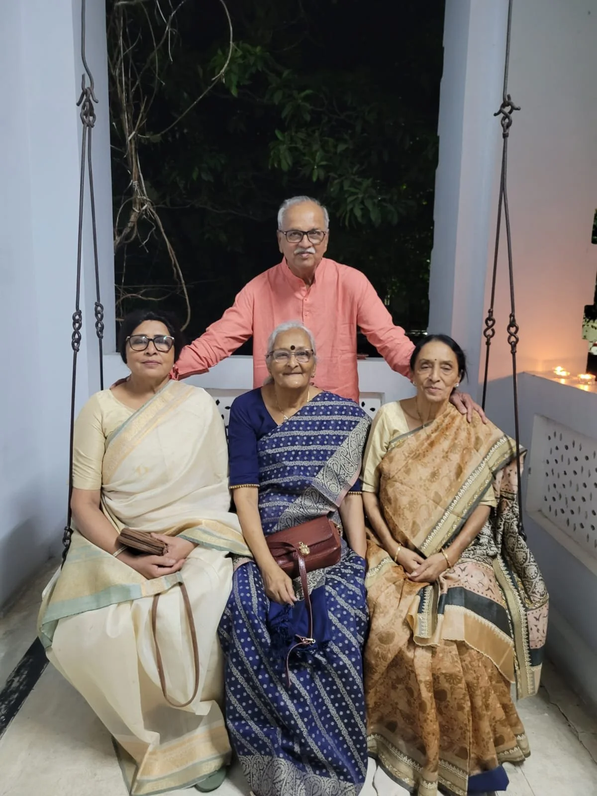 A group of five people, three women and two men, dressed in traditional Indian attire, sitting and standing on a swing on a porch at night with greenery in the background.