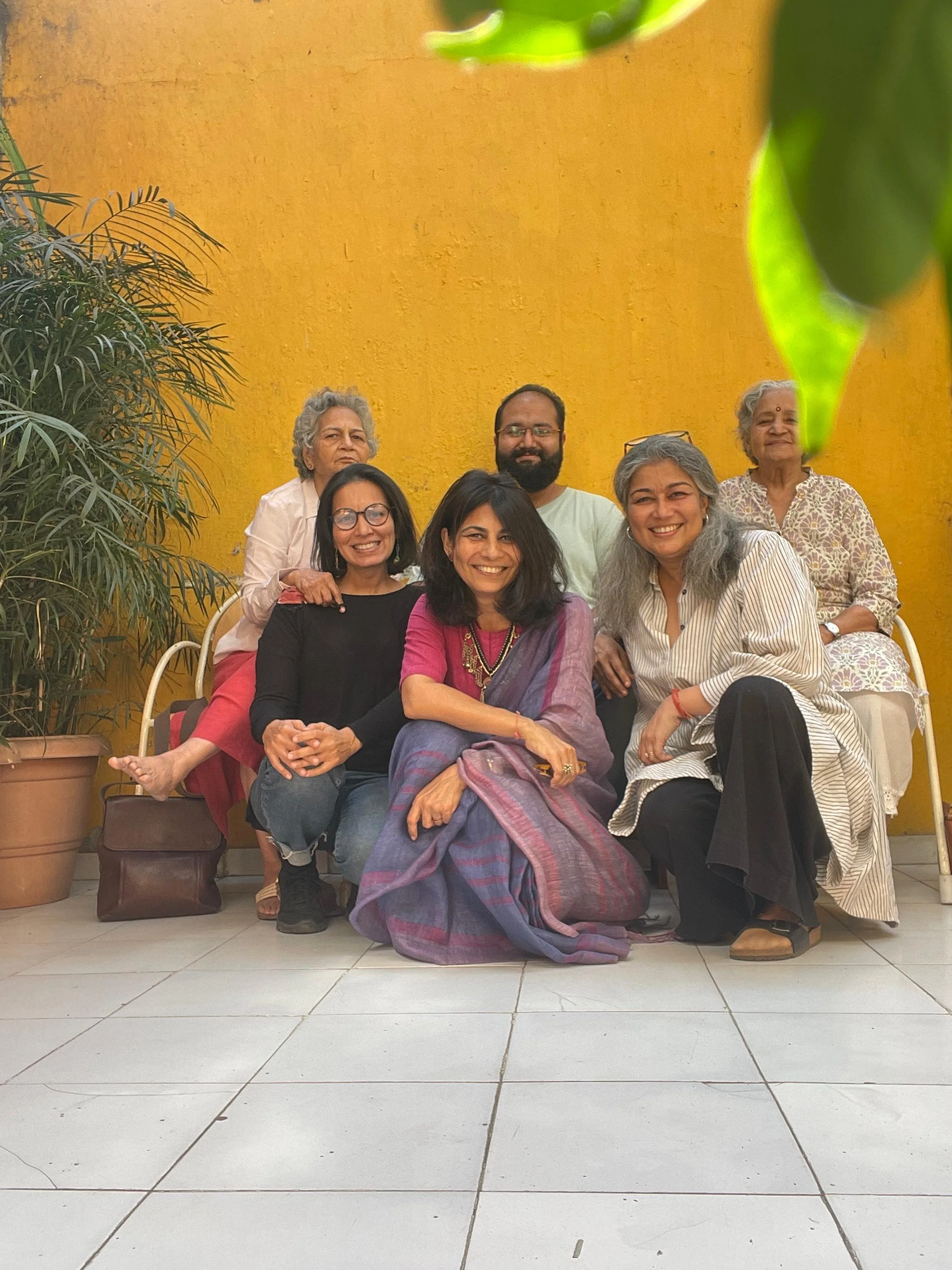 Group of six people, five women and one man, sitting and standing in front of a yellow wall, smiling at the camera, with potted plants on either side.
