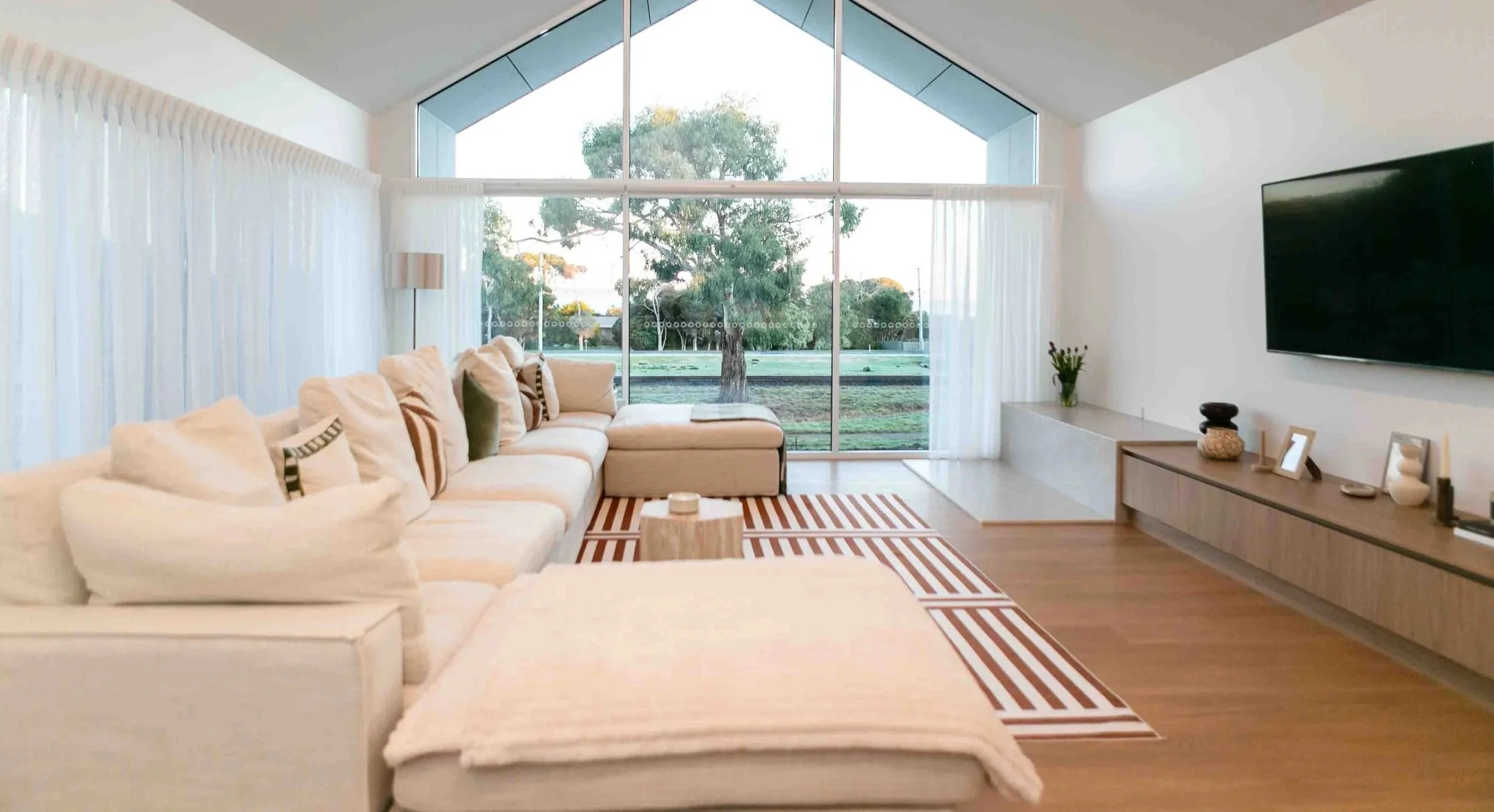 A stylish, modern living room with a comfortable couch and a pitched windoe wall with a view of a paddock and large gum tree. The home was built by MCC Projects in Leith, Tasmania.