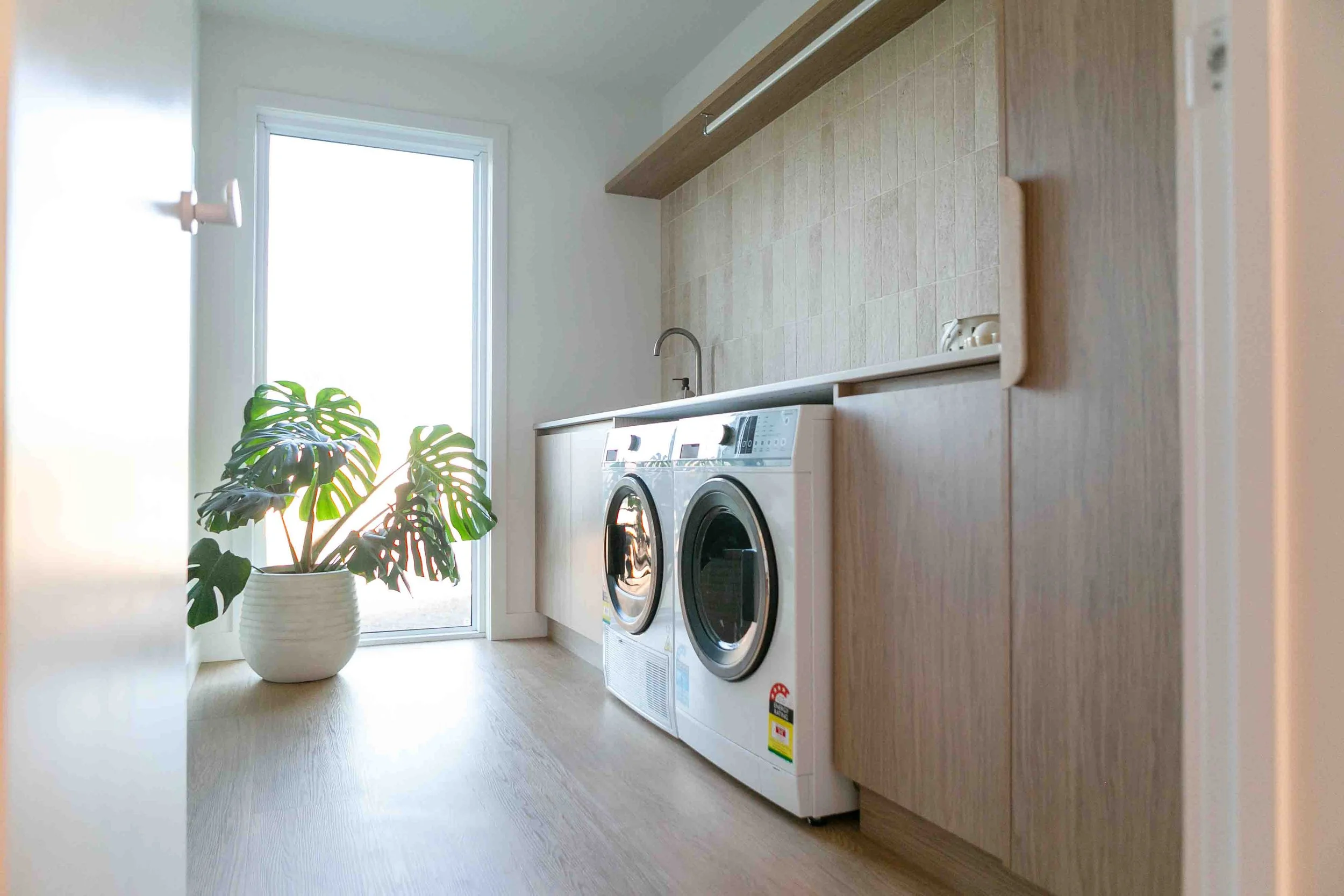 A well-lit laundry room showcasing a washer, dryer, sink, and tiled surfaces, with sunlight streaming through a window.