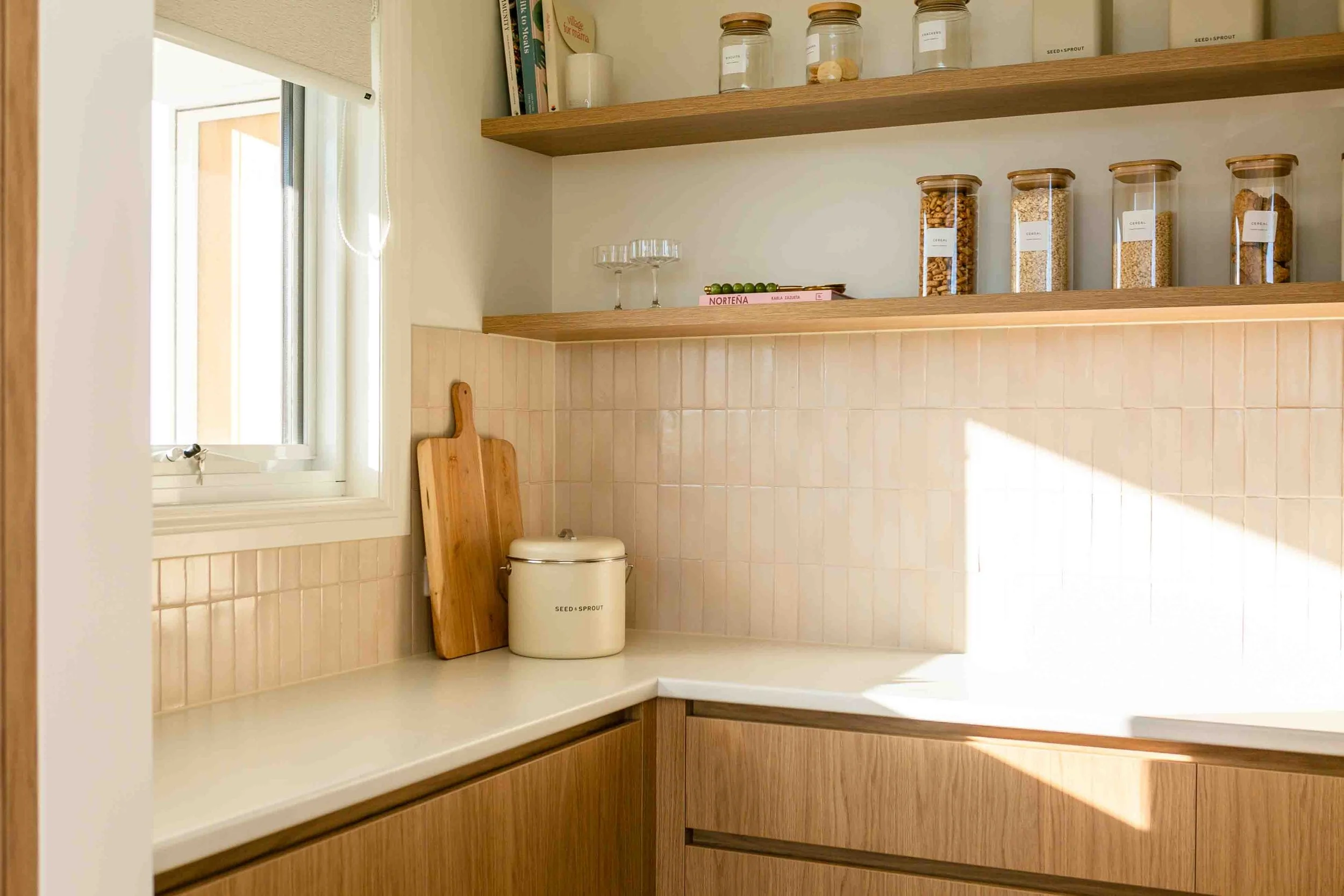 A stylish butler's pantry with pink tiles, floating shelves holding cookbooks and grains and a chopping board on the bench