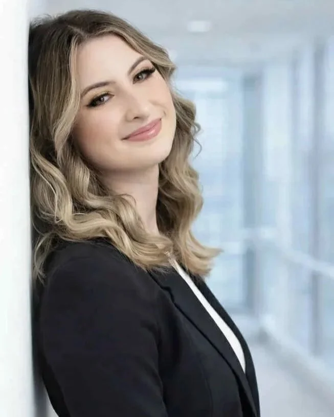 Young woman with wavy blonde hair and a black blazer, smiling and leaning against a wall in a modern building.