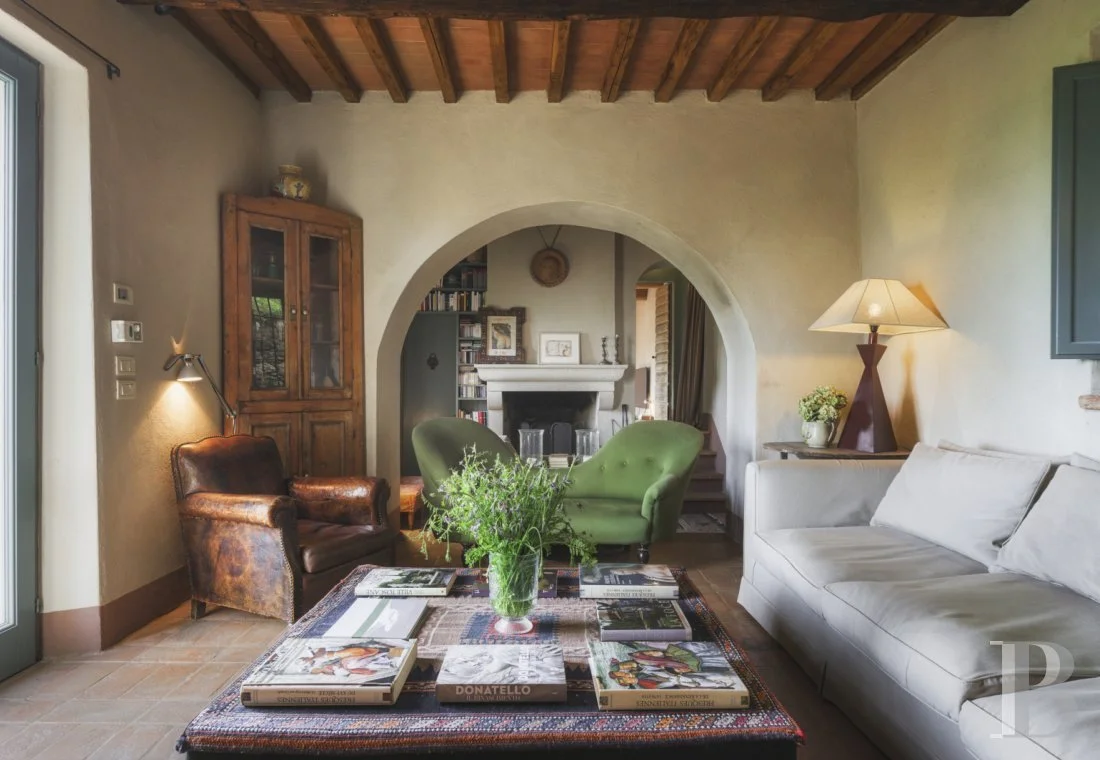 Salon avec canapé blanc, fauteuil en cuir, deux fauteuils verts, lampe, étagère en bois, cheminée, table basse avec livres et bouquet de fleurs.