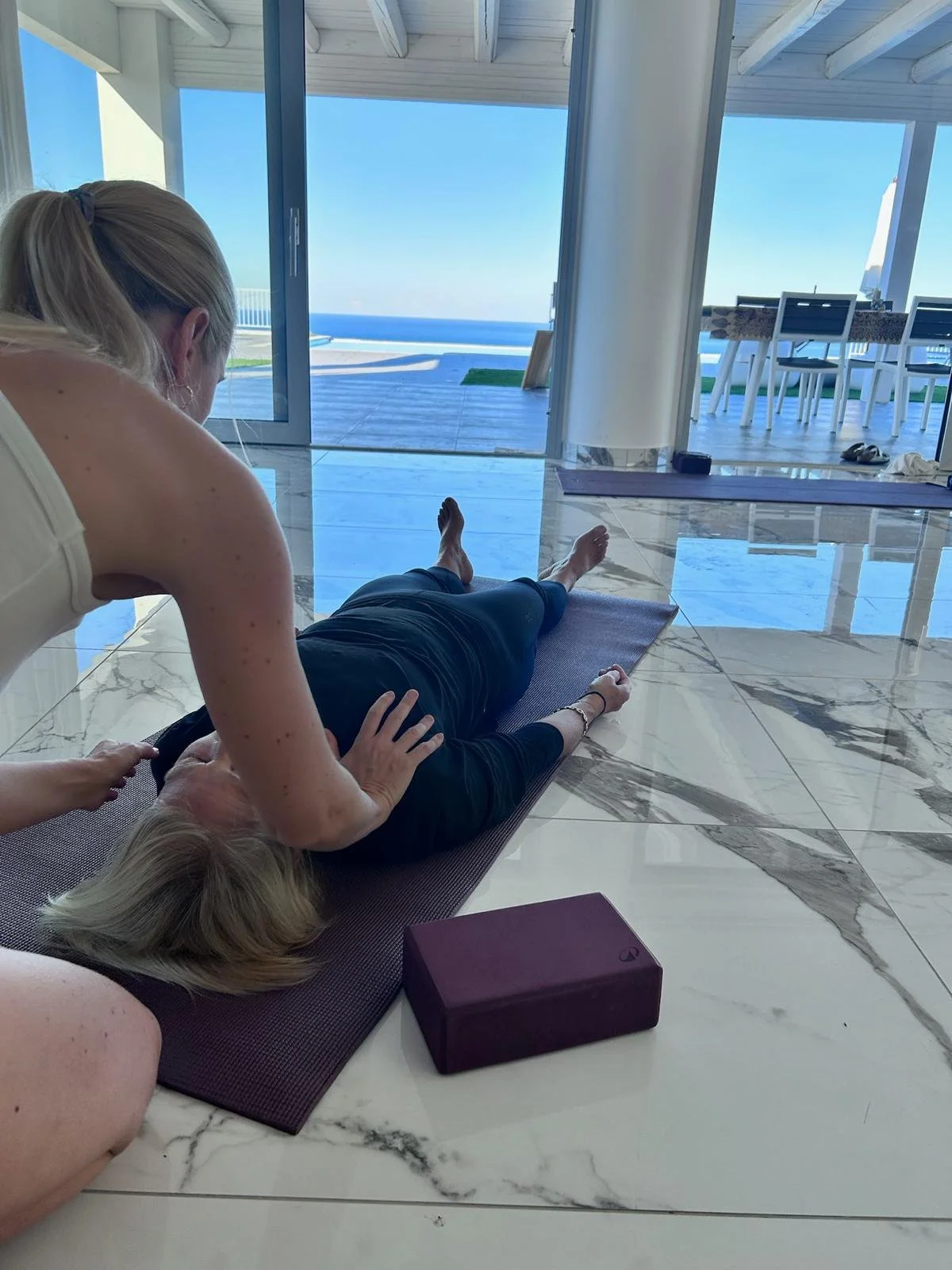 Une femme en position de relaxation avec une instructrice de yoga ou de massage dans une pièce lumineuse avec vue sur la mer.