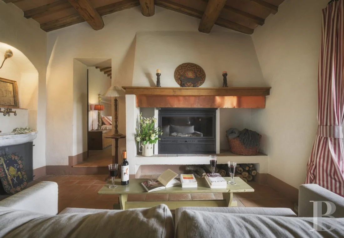 Salon avec cheminée en pierre, table basse avec livres, verres de vin, et une bouteille, canapé blanc, rideaux rayés rouge et blanc, plafond en bois avec poutres apparentes, lampe de lecture posée sur une cheminée en pierre, décor rustique.