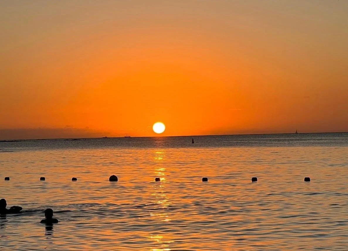 Coucher de soleil sur la mer avec des bateaux en silhouette à l'horizon, reflet du soleil sur l'eau, quelques personnes dans l'eau en premier plan.