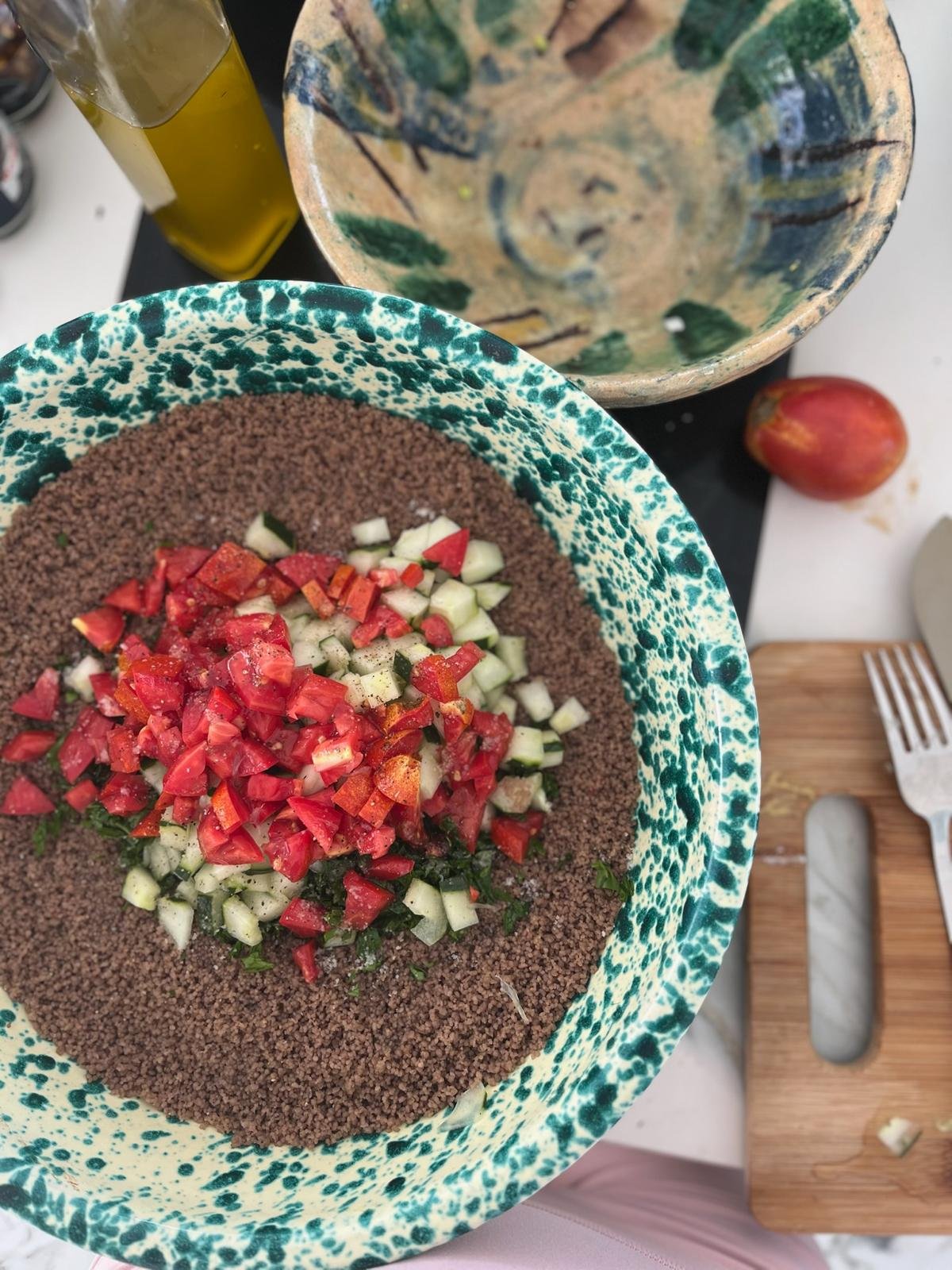 Une assiette de tabbouleh avec du boulgour, des tomates, du concombre, et du persil. À côté, une bouteille d'huile d'olive, une coupe, un fruit rouge, et une planche à découper avec un couteau et des morceaux de légumes.