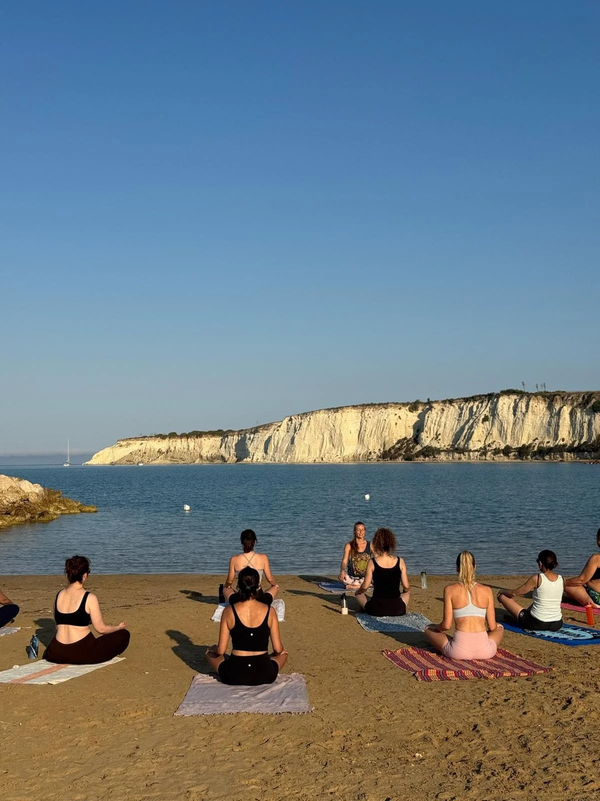 Groupe de personnes pratiquant le yoga sur la plage lors d'une séance en plein air, avec des falaises blanches et la mer en arrière-plan.