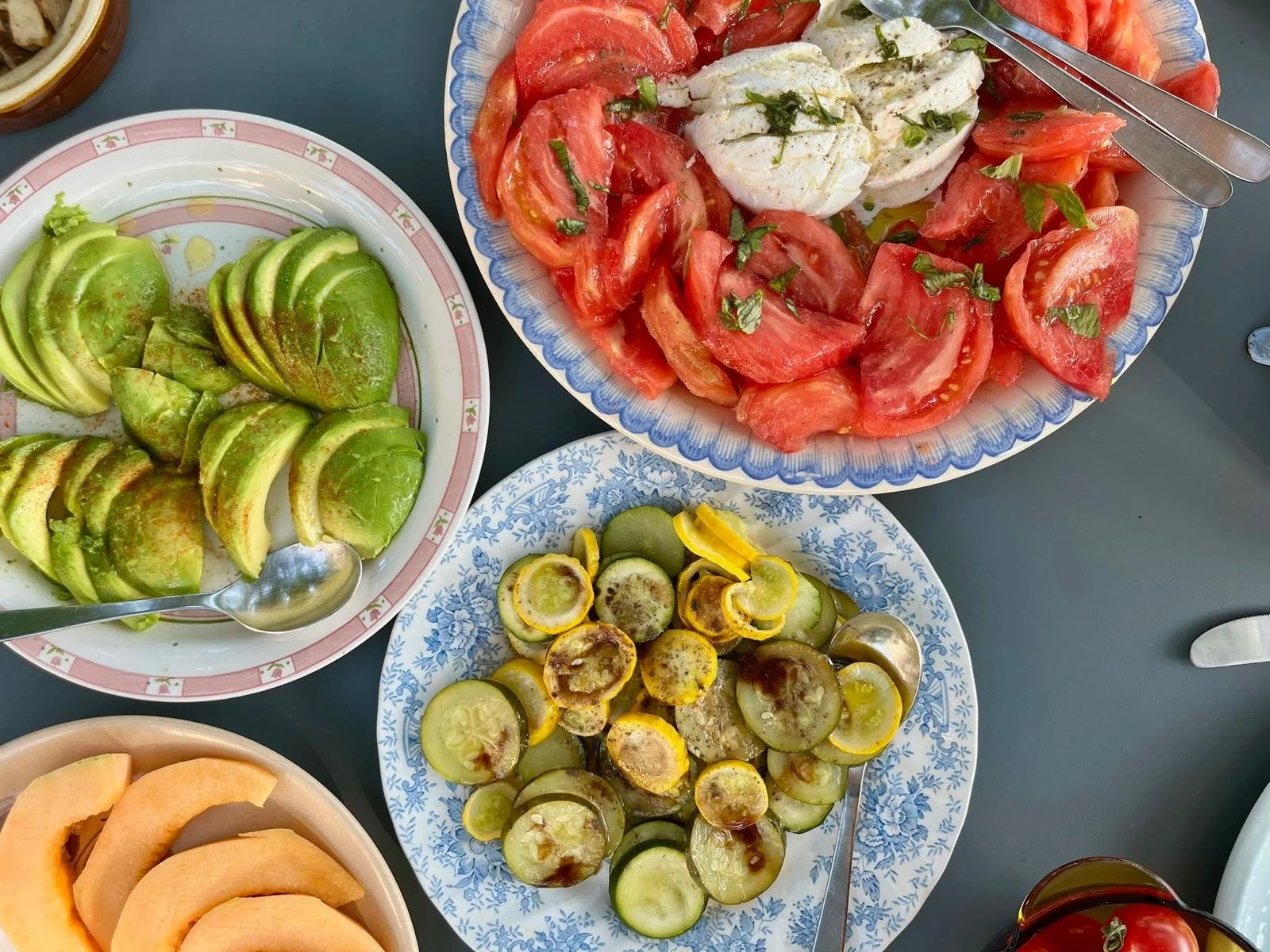 Plate de tranches de concombre, salade de tomates avec mozzarella, bol de melon tranché, et assiette de cornichons avec sauce dessus.