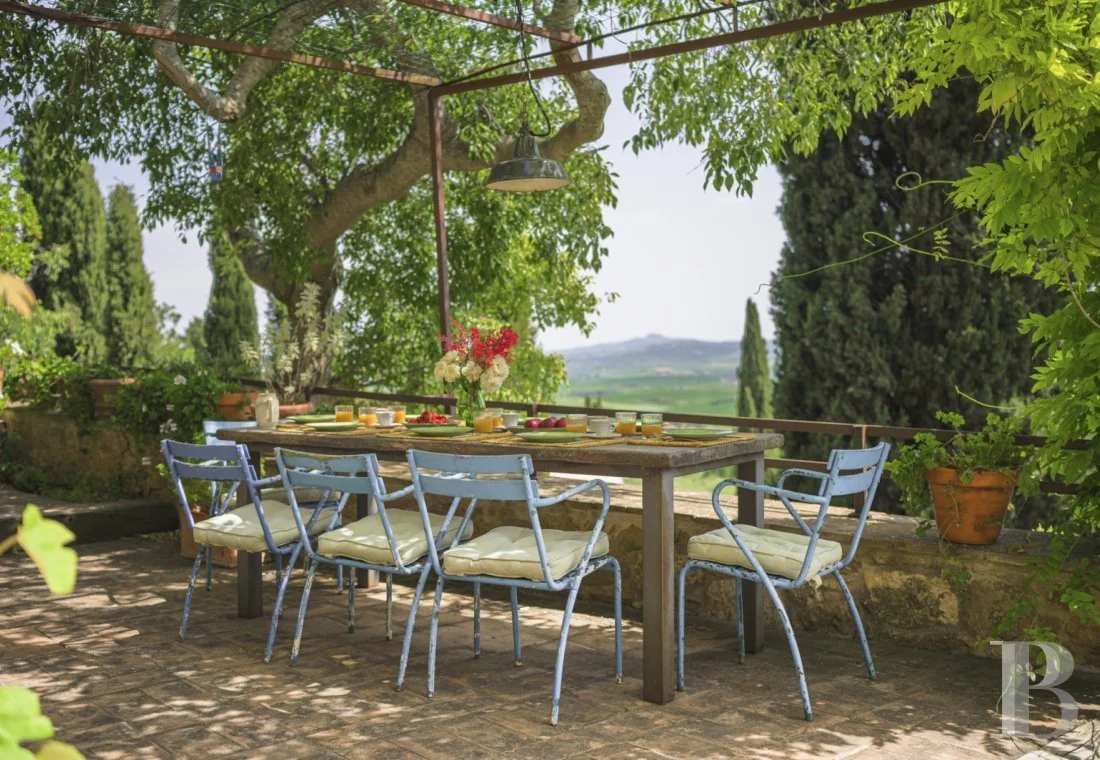Table de jardin en pierre avec chaises bleues sous un arbre, décorée pour un repas en plein air avec des fleurs et des boissons, dans un jardin verdoyant avec vue sur la campagne.