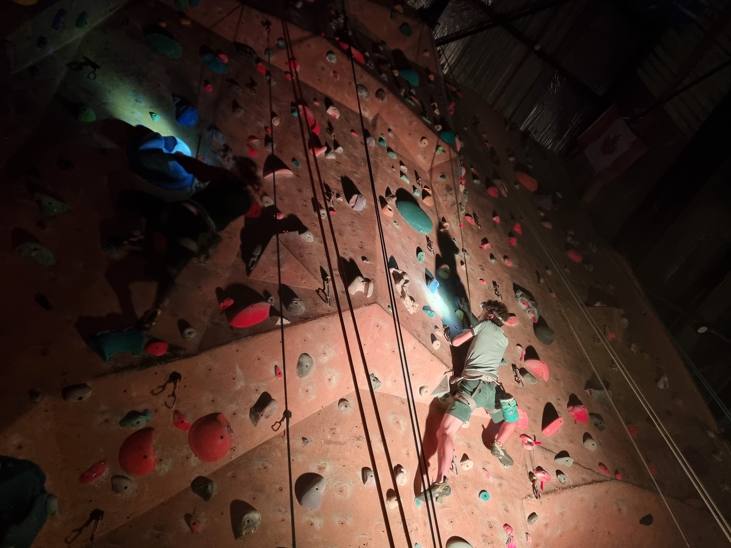 Person rock climbing indoors on a vertical climbing wall with various colored holds, wearing a harness and headlamp.