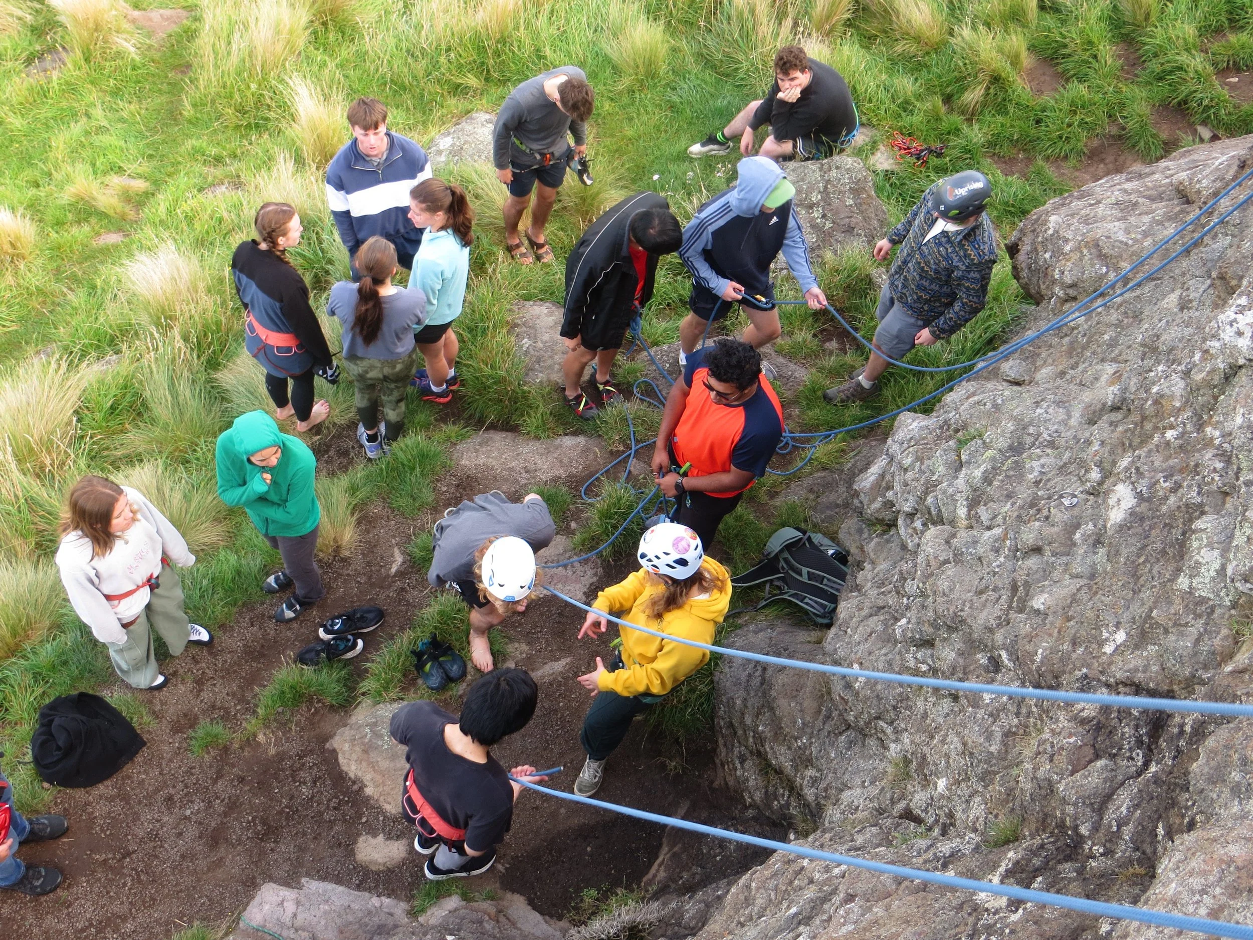 Group of people preparing to climb or rappel on a rocky outdoor terrain, some wearing helmets and harnesses, with grassy area surrounding the rocks.