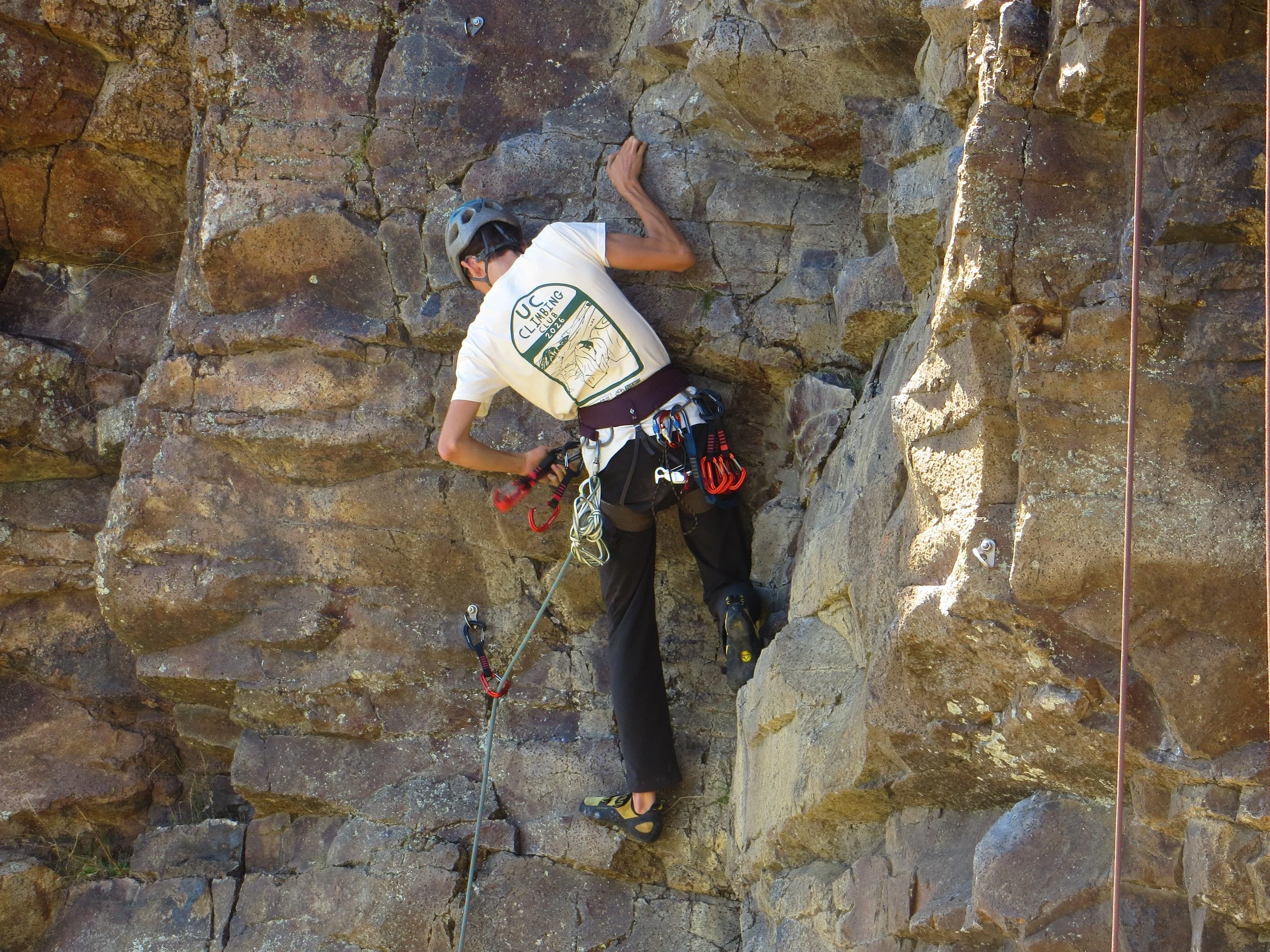 A man wearing a helmet and harness rock climbing on a vertical rock face outdoors.