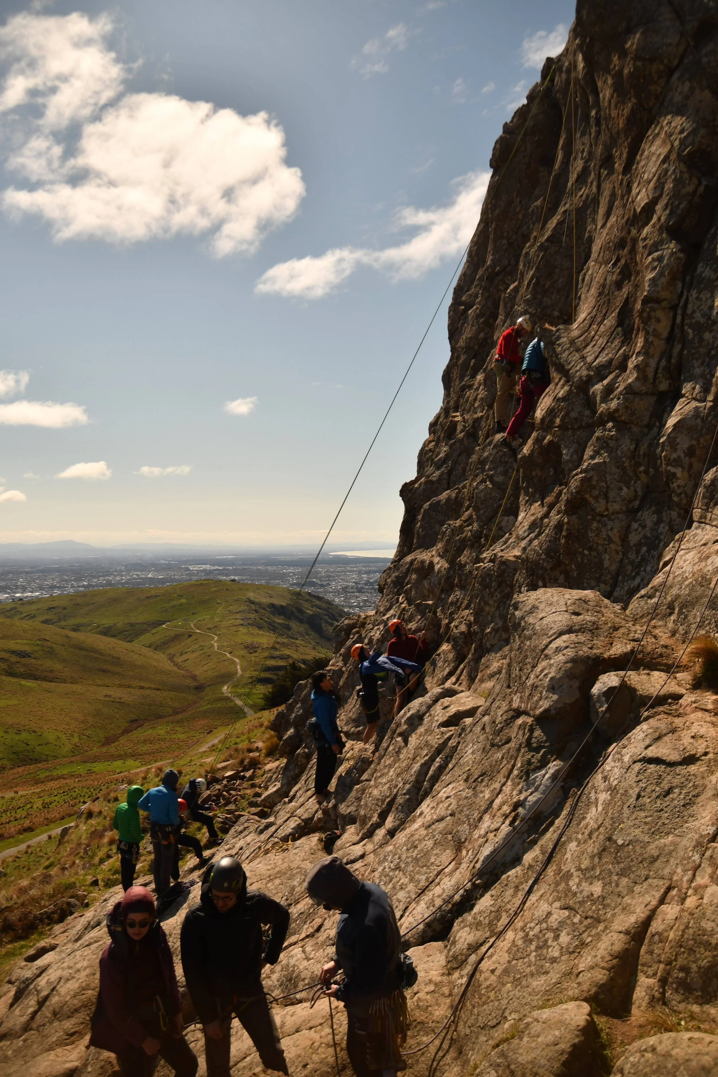 People rock climbing on a steep mountain with a trail and landscape visible in the background under a partly cloudy sky.