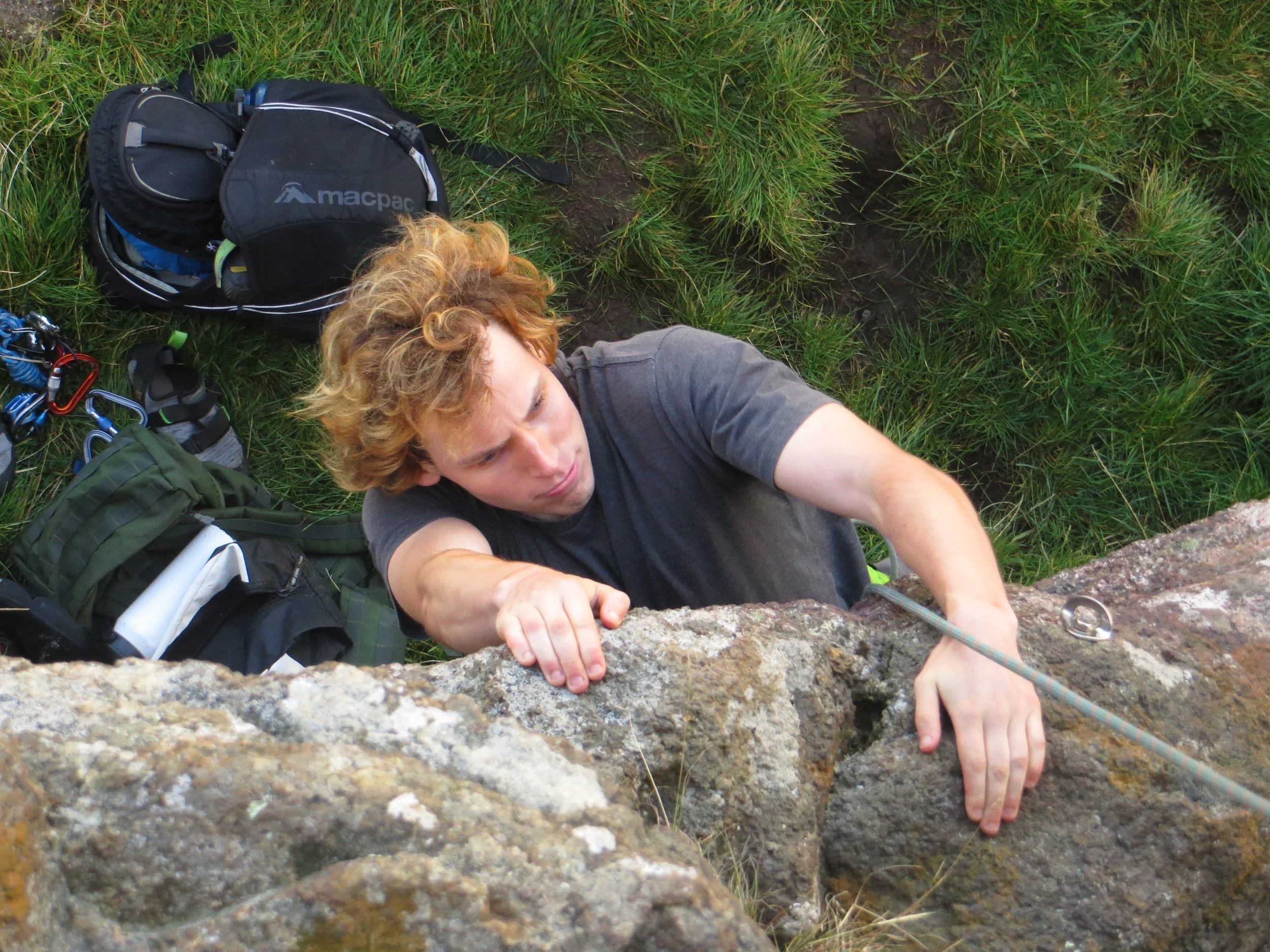 A young man rock climbing outdoors, gripping a rock with his right hand, surrounded by climbing gear and backpacks on green grass.