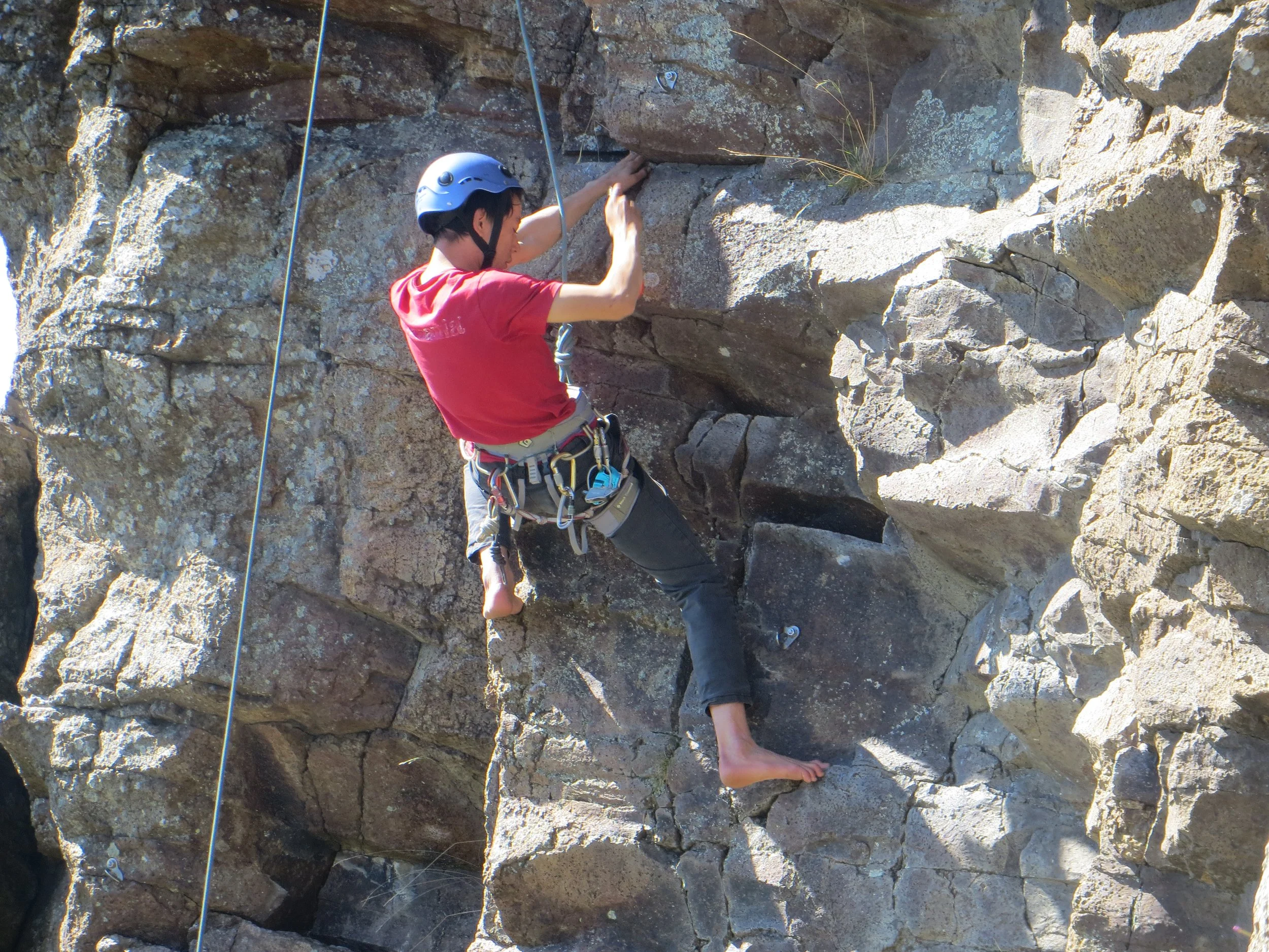 A person rock climbing on a vertical rock face outdoors, wearing a red shirt, climbing harness, and a helmet.