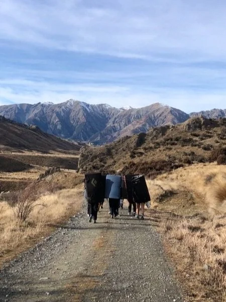 Group of people walking on a dirt trail in a mountainous landscape with dry grass and rugged terrain.