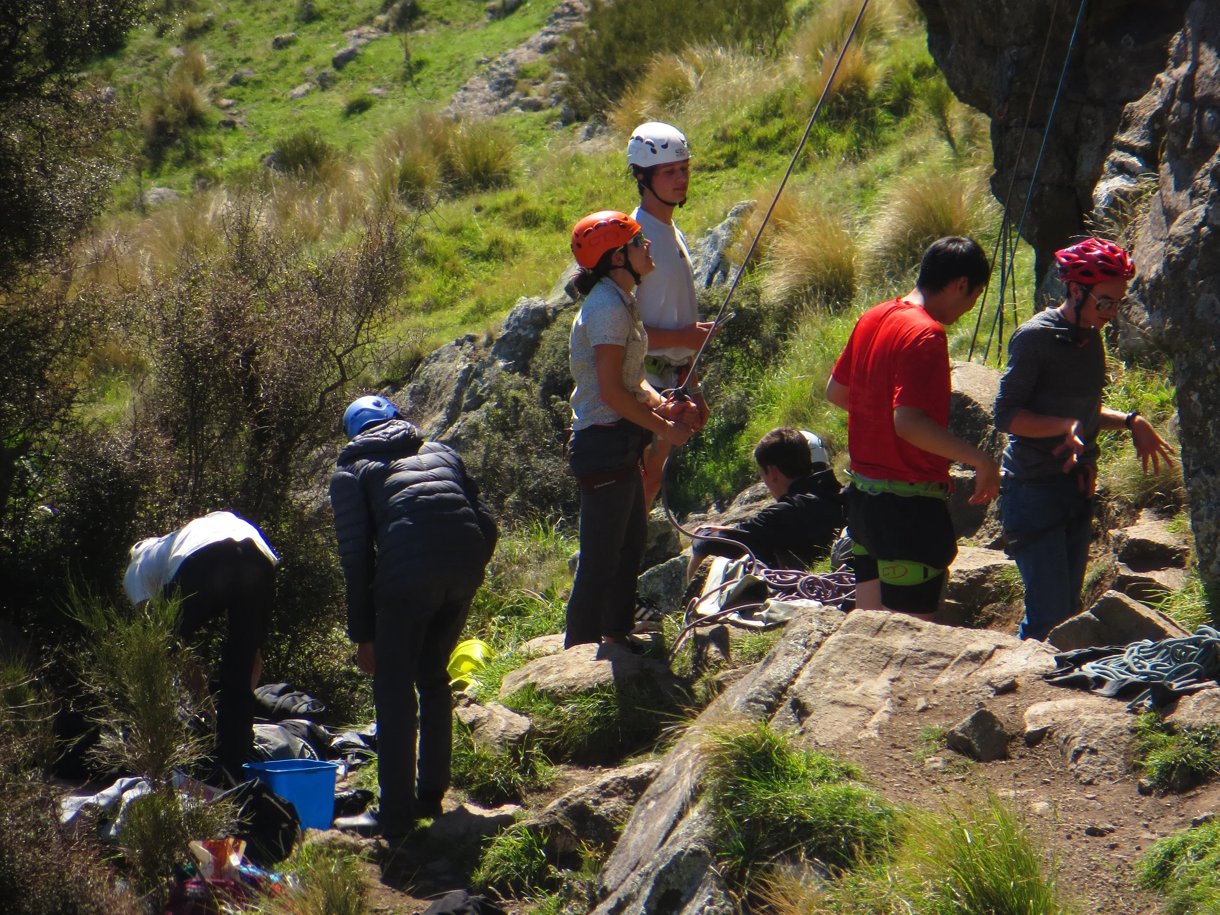 A group of people preparing for rock climbing or rappelling outdoors on a rocky hillside, wearing helmets and harnesses.