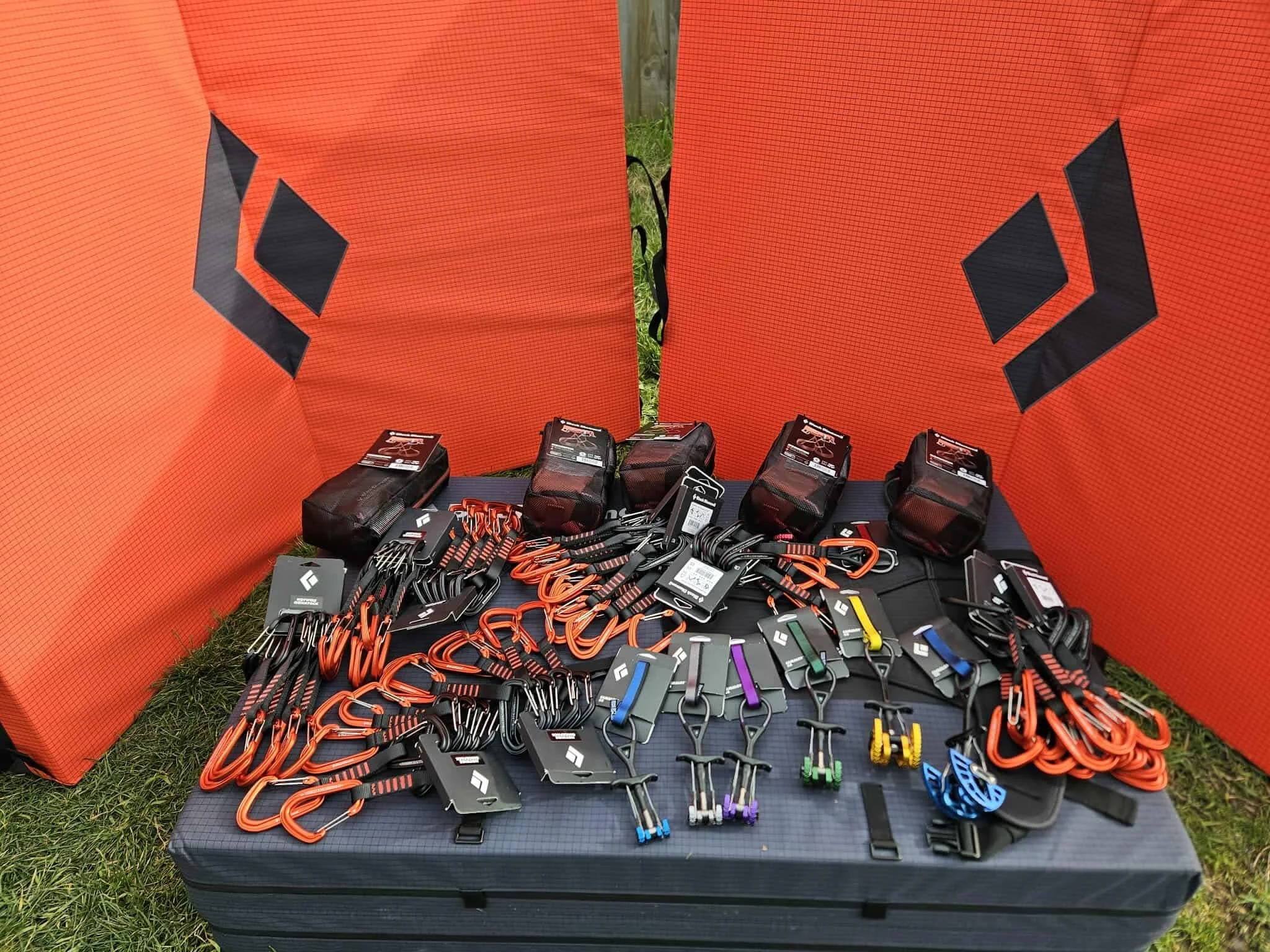 Climbing gear including carabiners, harnesses, and chalk bags spread on a suitcase with orange crash pads in the background.