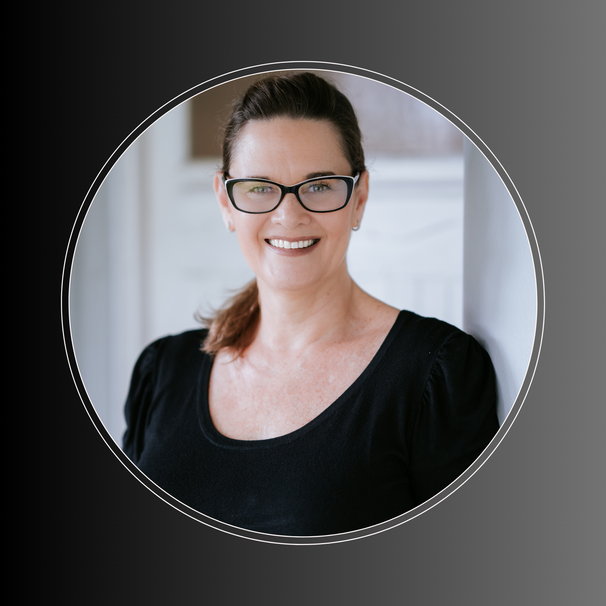 Smiling woman with glasses, wearing a black top, leaning against a wall indoors.